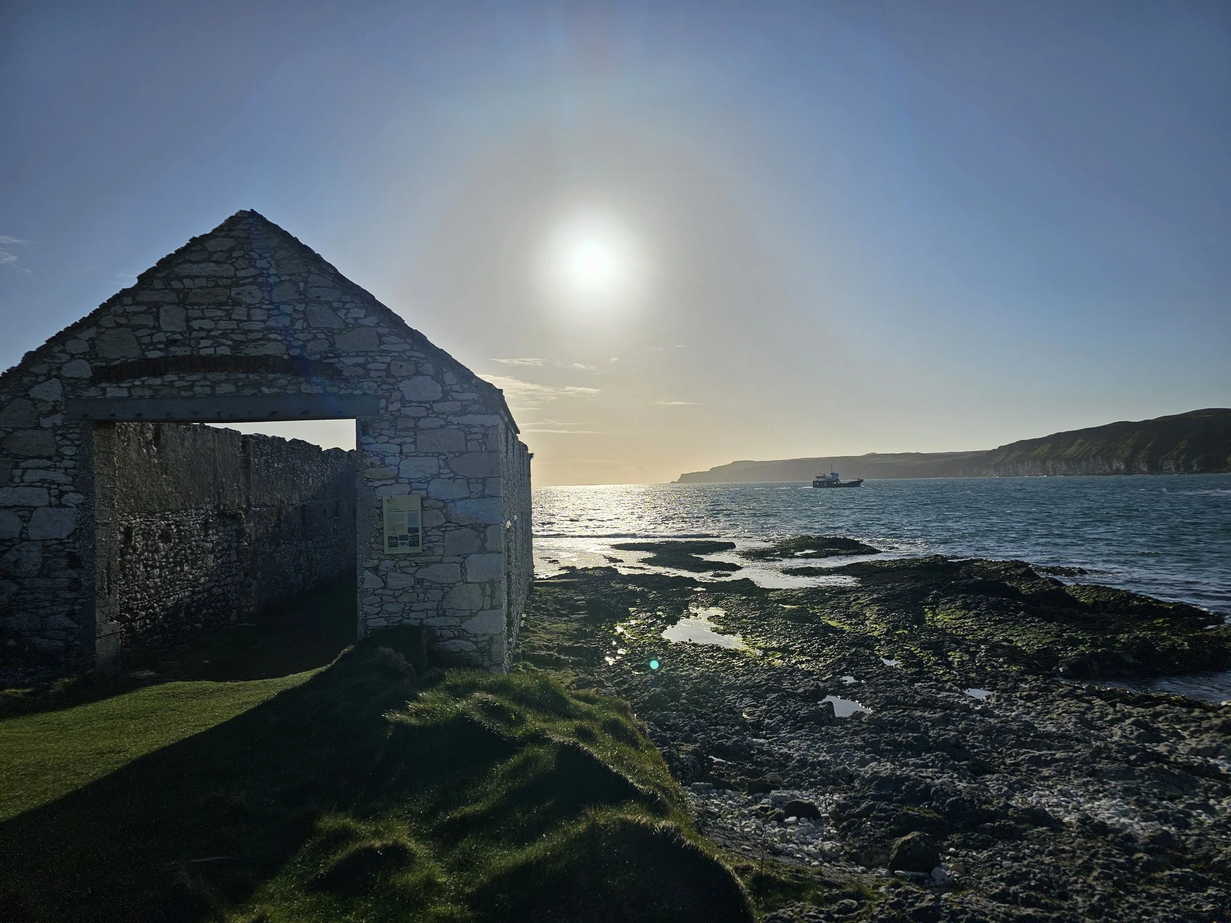 A stone building with an archway stands on a grassy shoreline, with a large body of water and cliffs in the background. The sun is low in the sky, creating a silhouette effect and a bright glare on the water.