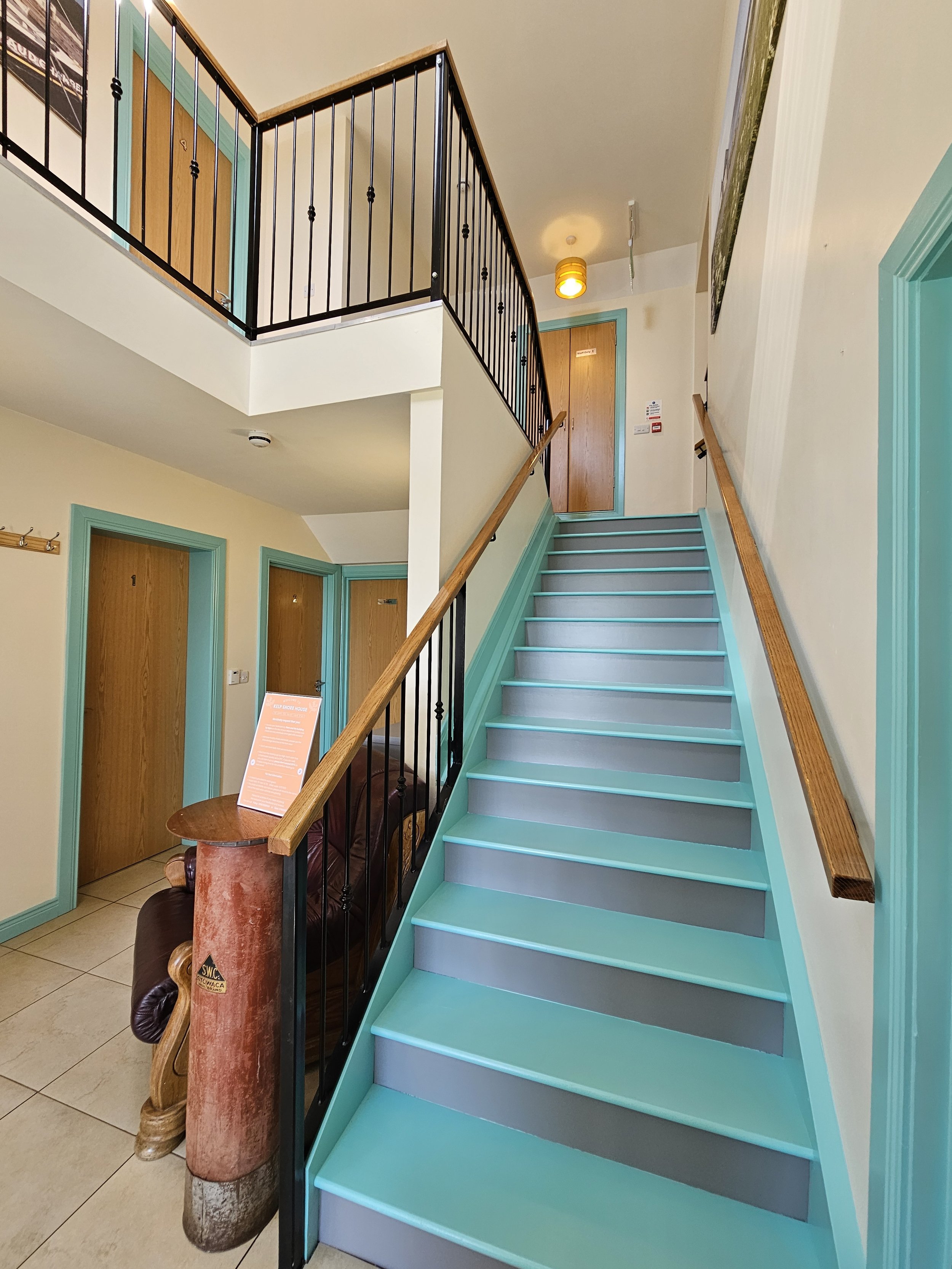 Indoor staircase with teal and gray steps, wooden handrail, and black metal railing. Wooden doors with teal trim on the upper floor and below. A yellow ceiling light, a small wooden table with a chair, and a staircase connecting two floors.