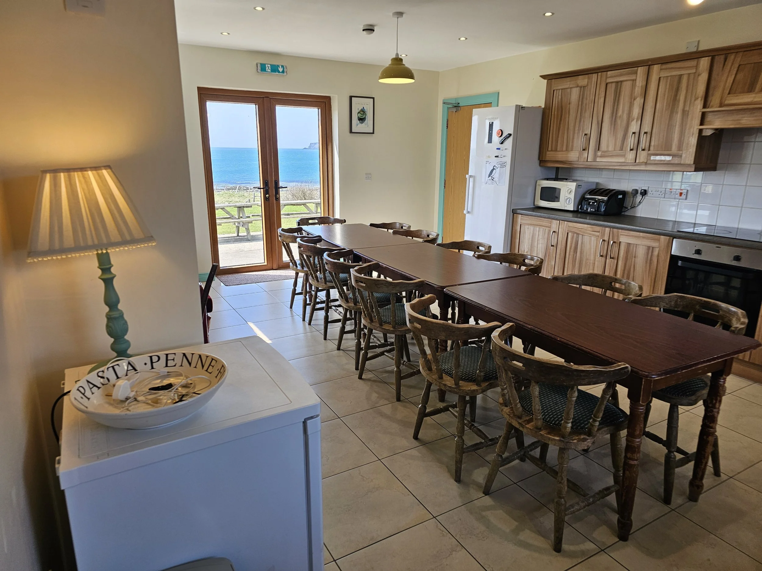 Inside view of a kitchen and dining area with a view of the ocean through glass double doors, wooden dining table with chairs, wooden kitchen cabinets, microwave, toaster, and a white freezer with a decorative bowl on top.
