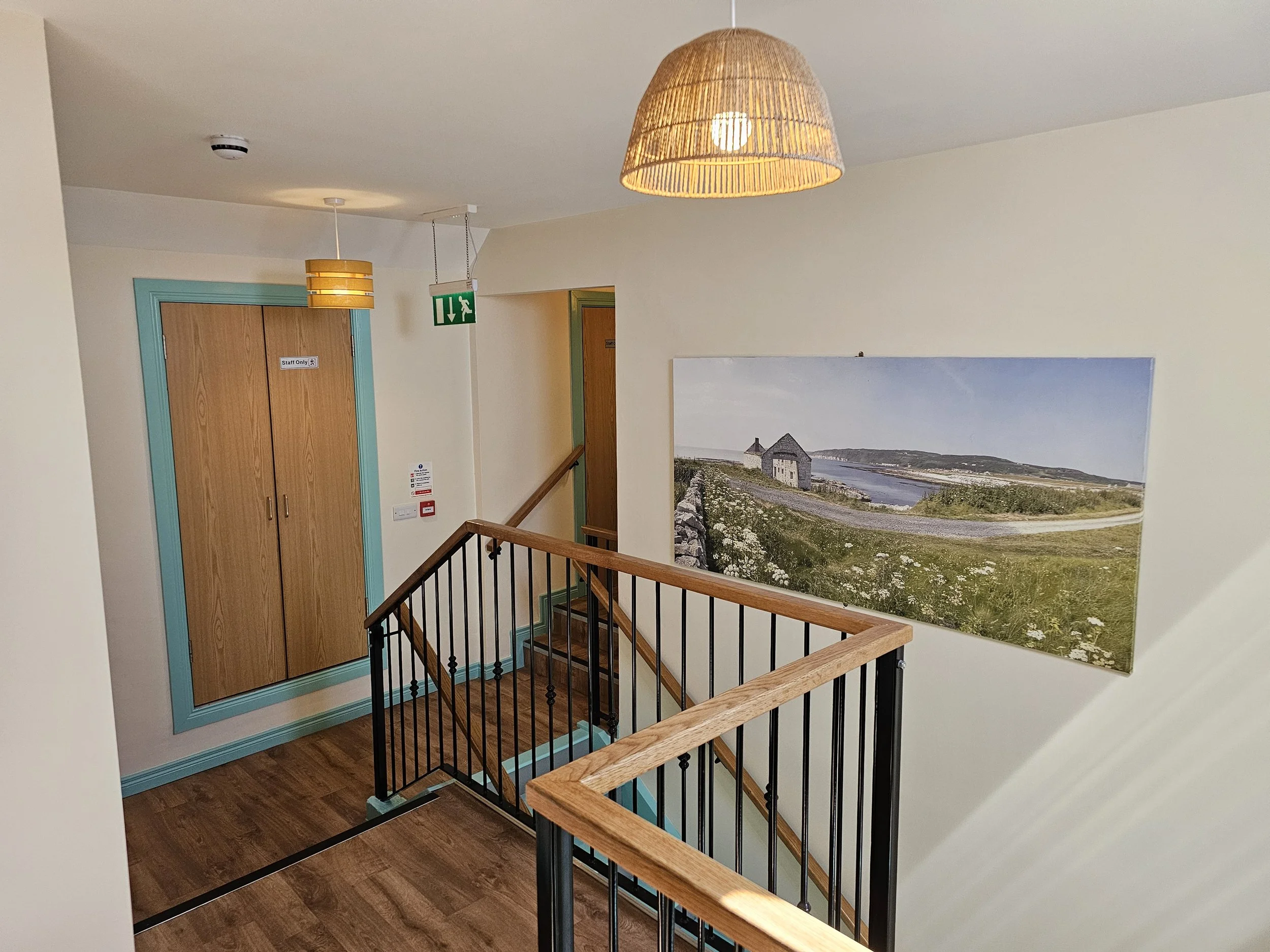 Interior view of a stairwell landing with wooden flooring, a railing with wooden top and black metal spindles, a large wall art depicting a landscape with a river, house, and mountain, a wooden door with a sign, and ceiling lights, one with a wicker 