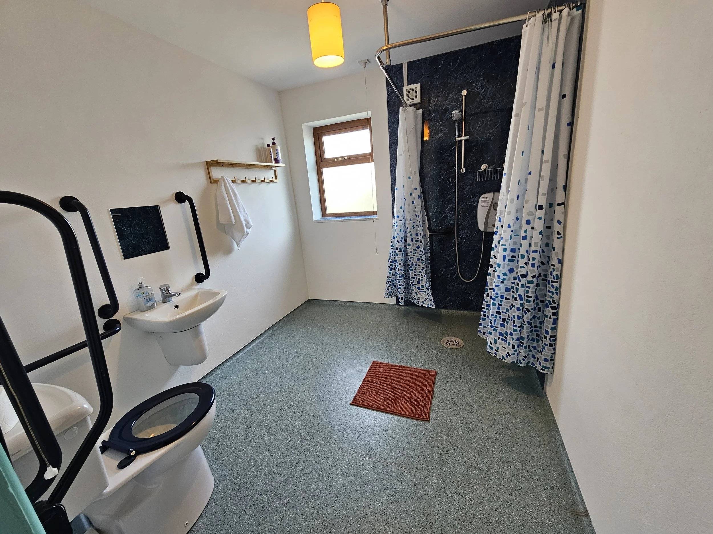 A bathroom with a white sink, a mirror, black grab bars, a small window, a shower with a curtain, a red rug, and various bottles on a wall shelf.