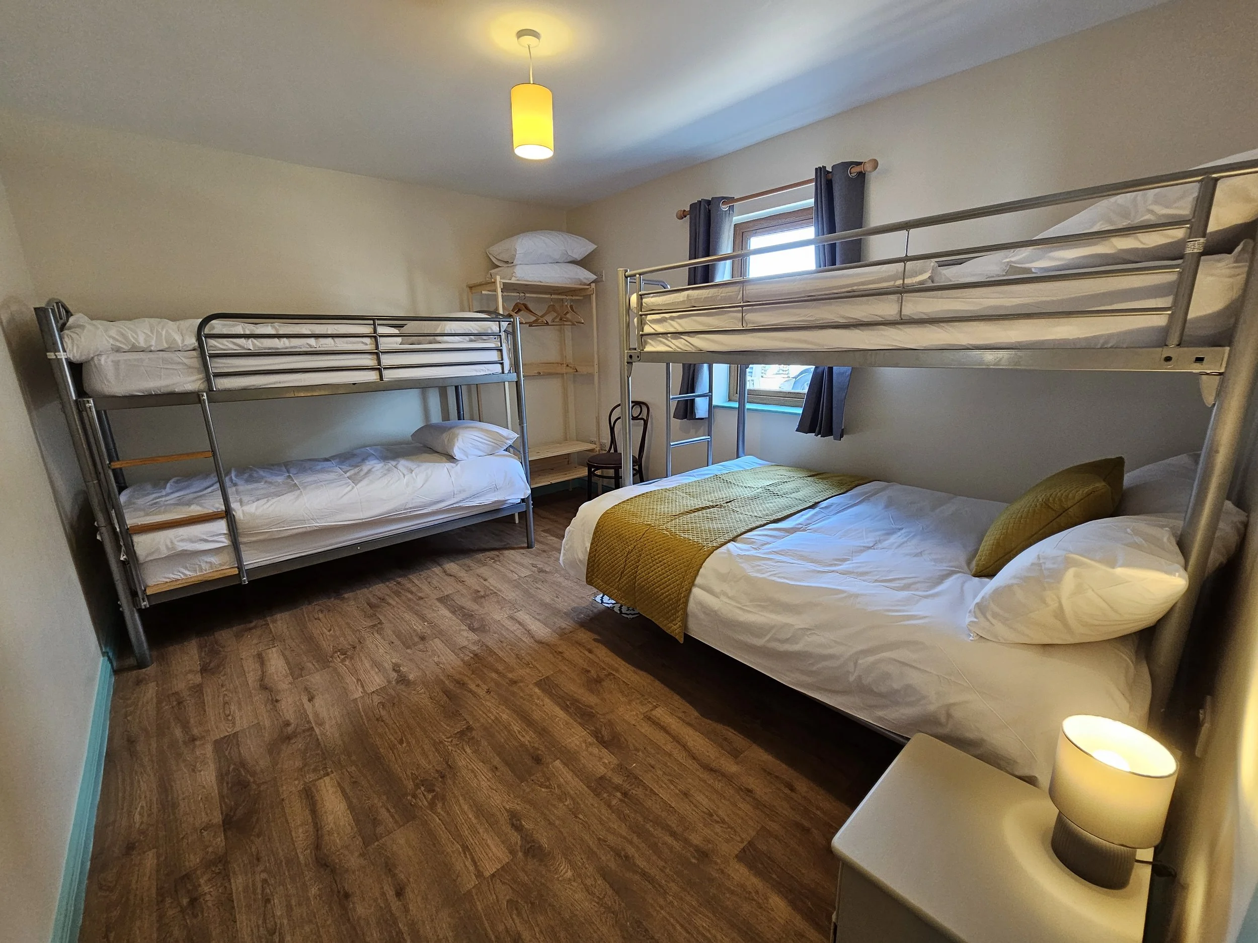 A bedroom with two metal bunk beds, a window with dark gray curtains, a wooden shelf with pillows, a wooden chair, a small white nightstand with a beige lamp, wooden flooring, and a ceiling light with a yellow lampshade.