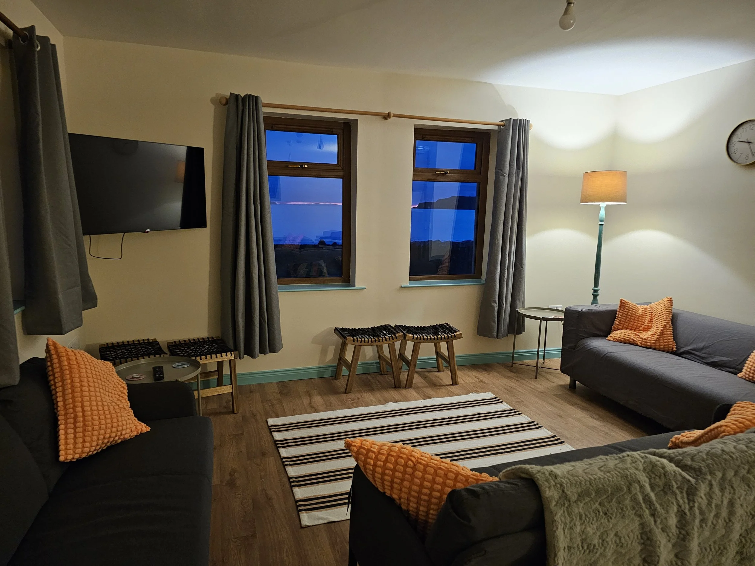 Living room with two sofas with orange and green textured pillows, a small round side table, striped rug, two wooden stools, two large windows with dark brown frames and gray curtains, a floor lamp, a wall clock, and a flat-screen TV on the wall. Out