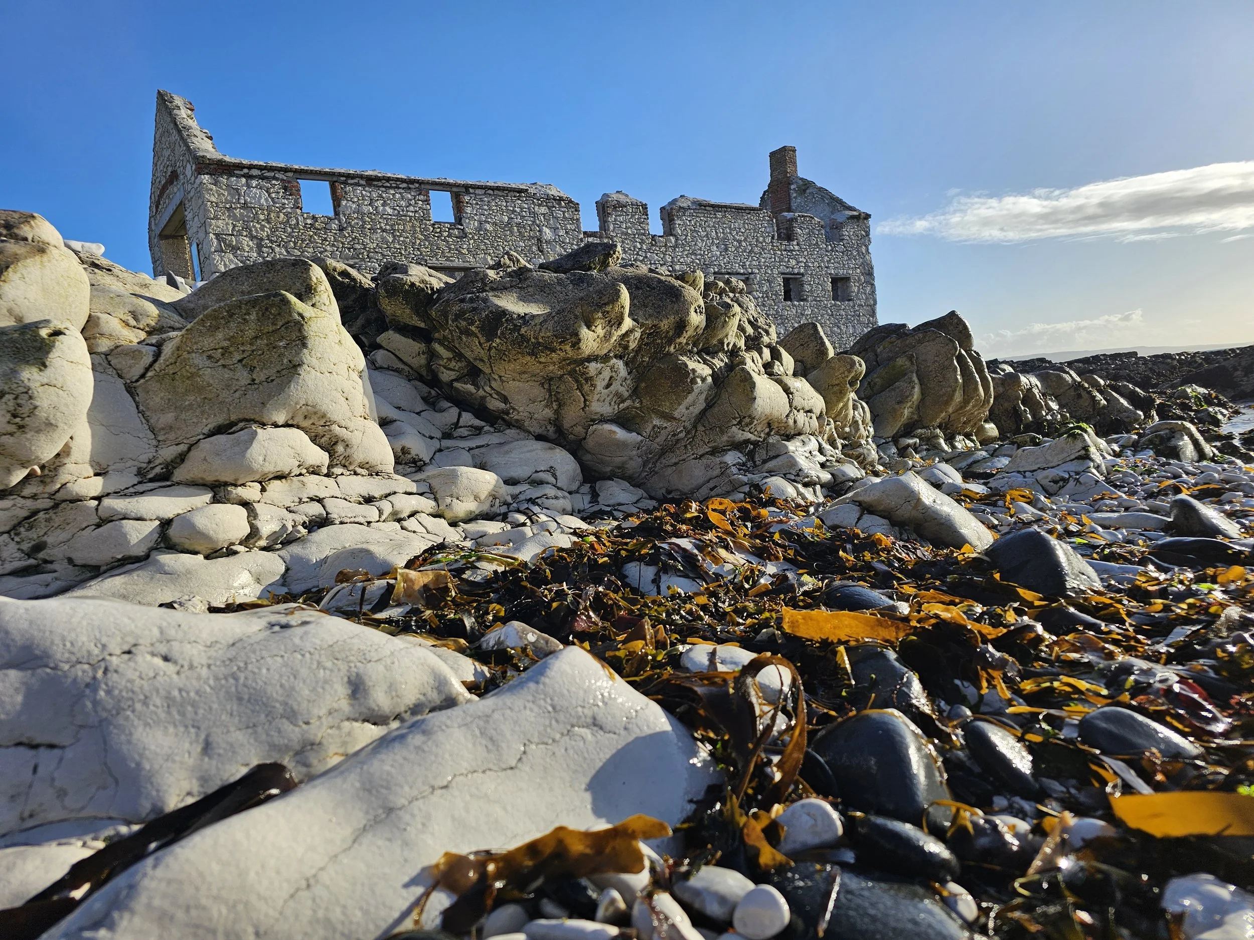 Ruined stone building on rocky shoreline with blue sky and clouds in the background.
