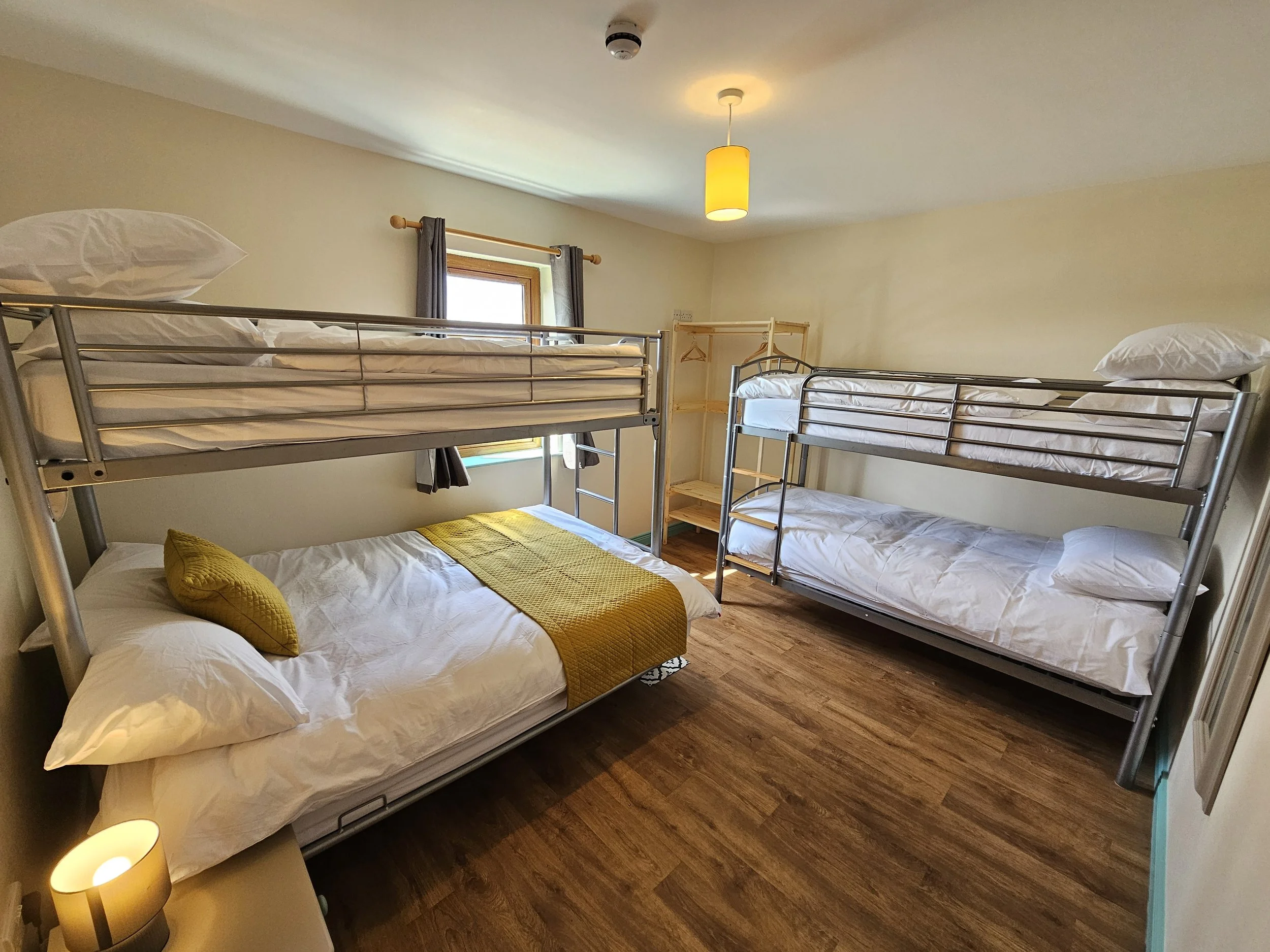 Room with four metal bunk beds, a window with gray curtains, a yellow ceiling light, and a yellow cushion on a bed, with wooden flooring.