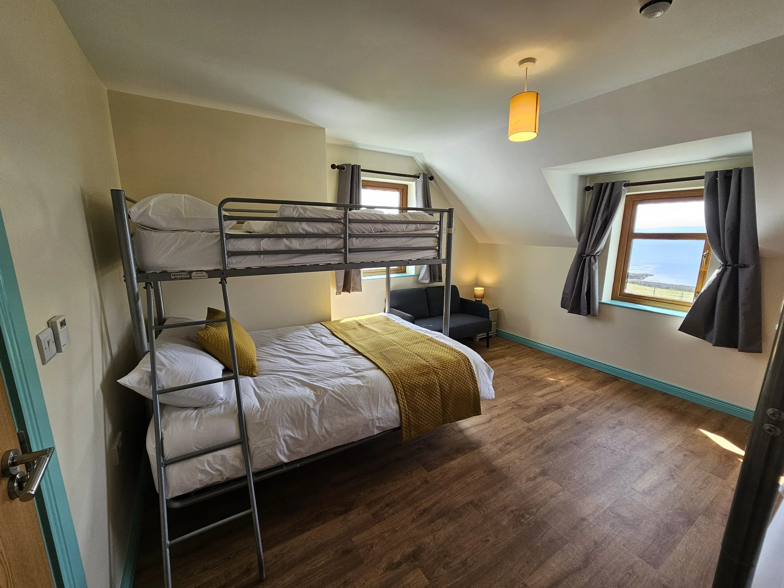 A bedroom with a bunk bed, a bed with a yellow blanket, a black sofa, two windows with gray curtains, a ceiling light, and wooden flooring.