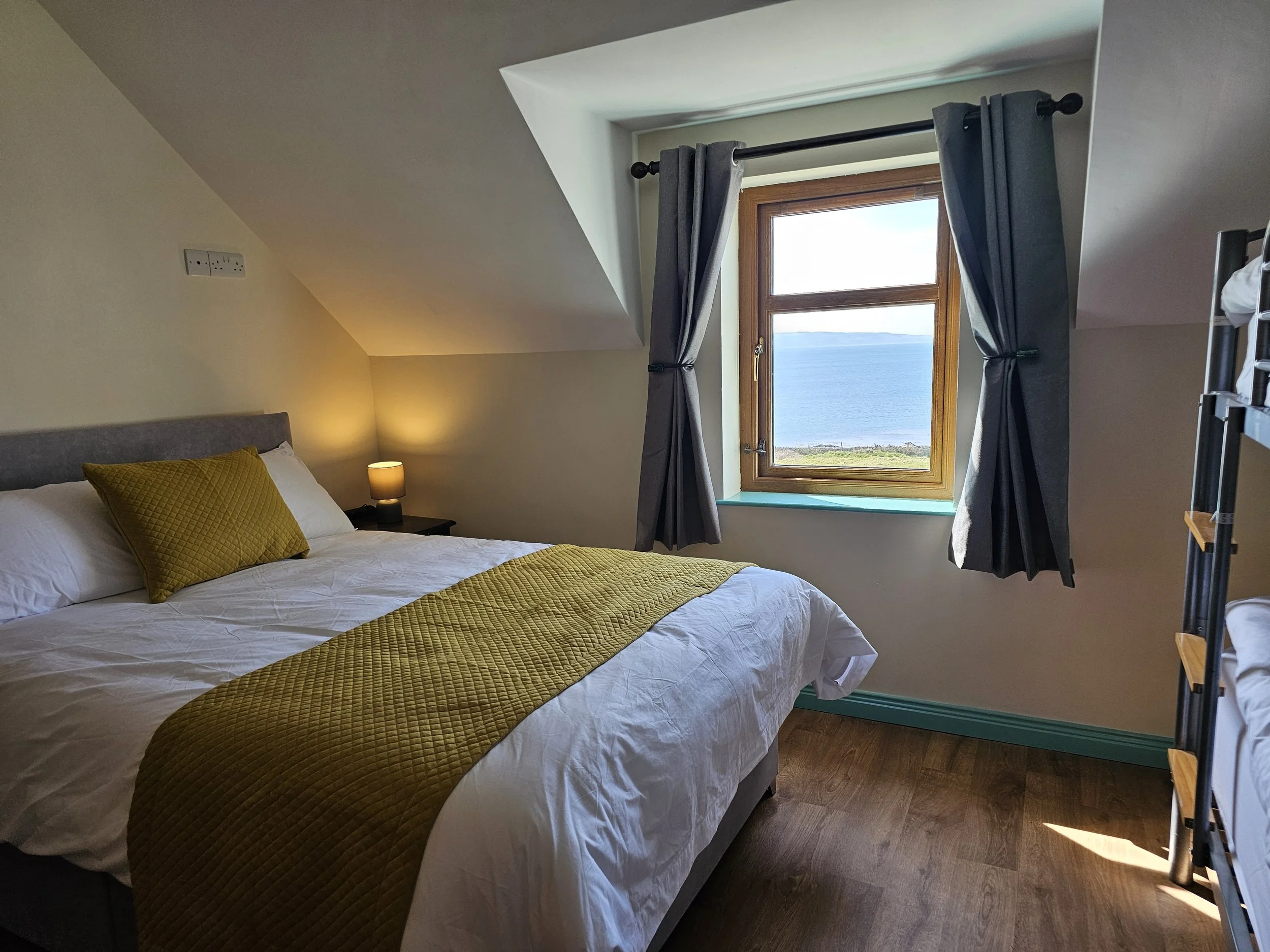 A cozy bedroom with a bed featuring white linens and a yellow quilted blanket and pillow. There is a small black bedside lamp on a nightstand next to the bed. A window with dark curtains and a scenic ocean view is above the bed, letting in natural li