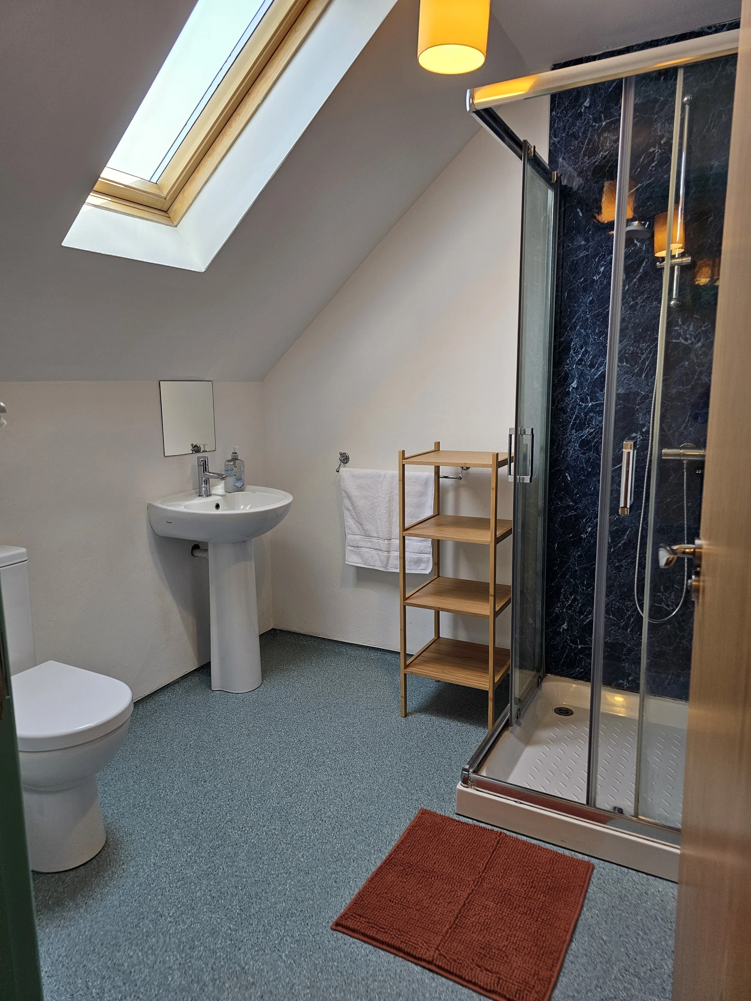 A bathroom with a skylight window, a white toilet, a pedestal sink with soap dispenser, a mirror, a wooden shelf with a towel, a shower with black marble walls, and a red rug on blue carpeted floor.