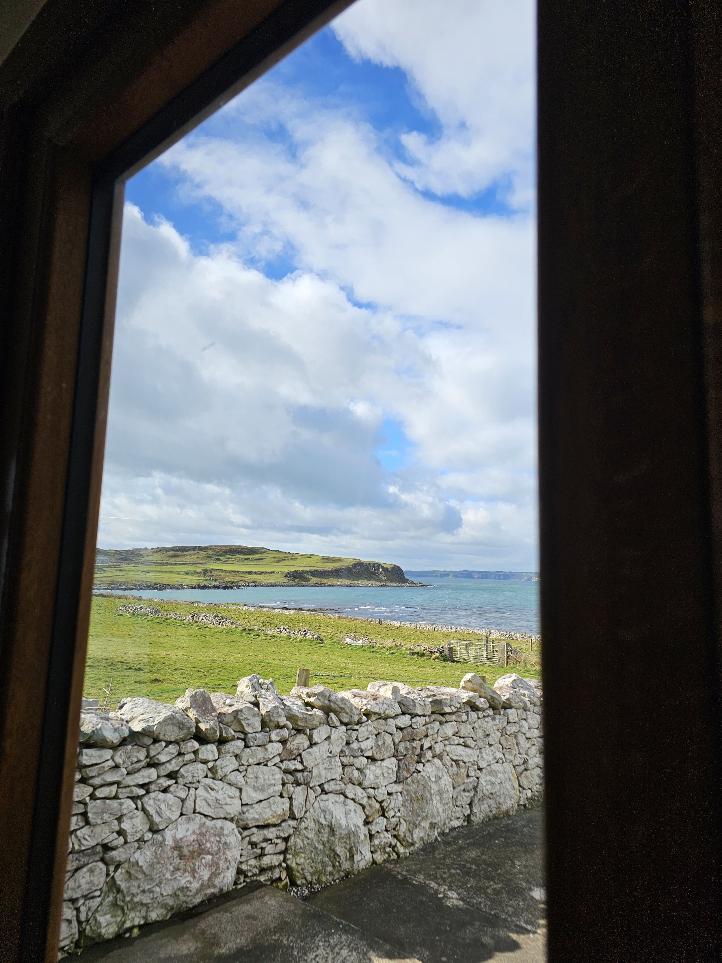 View of a coastline with green fields, a rocky stone wall, and a body of water, seen through a window with a black frame.