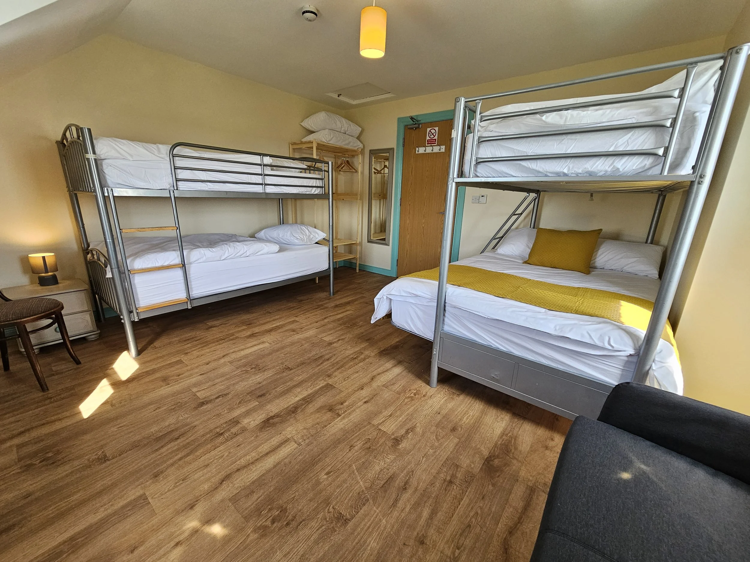 Hotel room with two sets of bunk beds, a wooden bedside table with a small lamp, a small chair, a wooden shelving unit with pillows, a mirror, and a door with a no smoking sign. The room has yellow walls, wooden flooring, and a ceiling light.