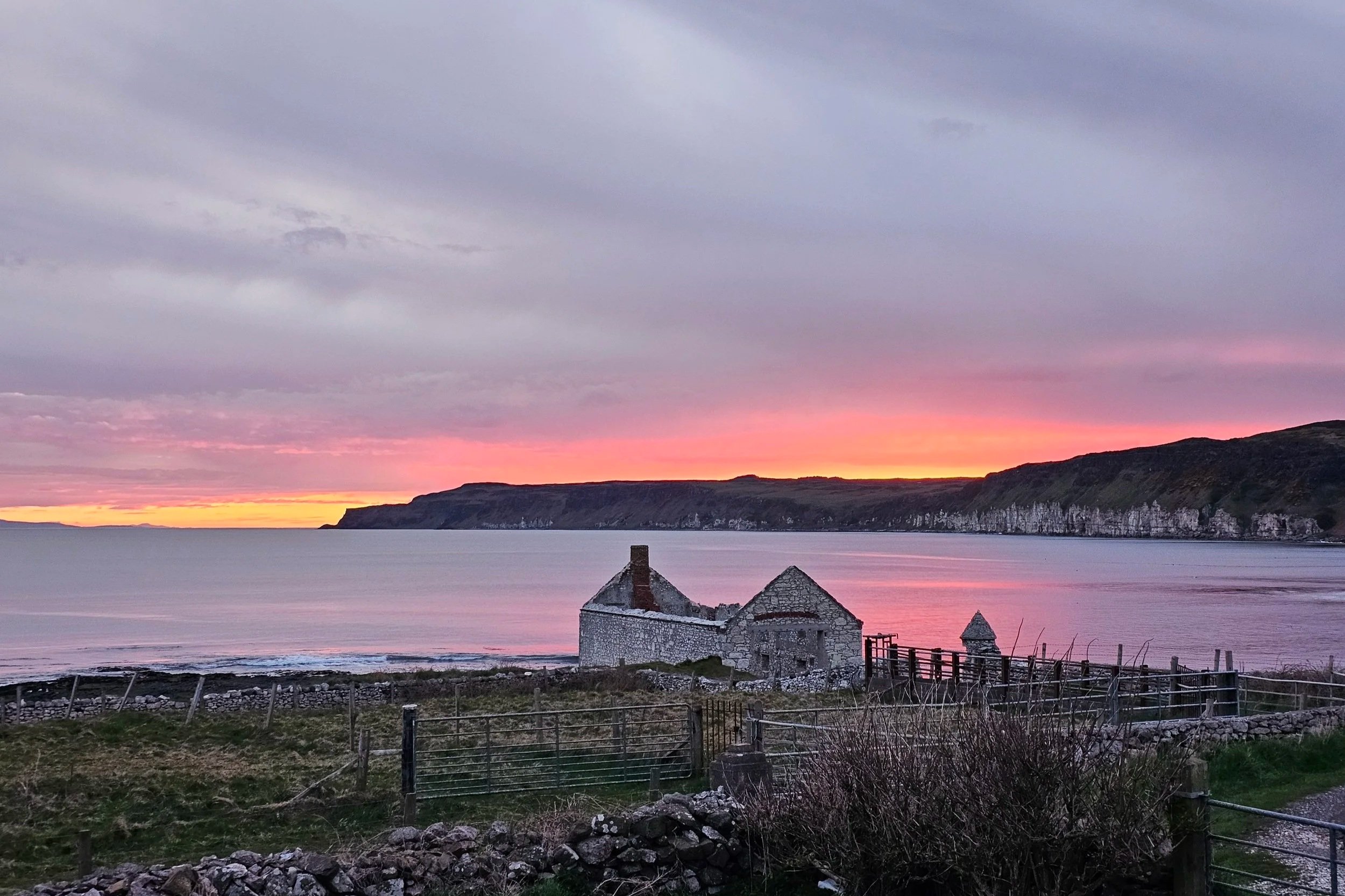 A scenic coastal landscape at sunset with a cloudy sky, distant cliffs along the shoreline, calm water, and rustic buildings with stone walls and tiled roofs in the foreground.