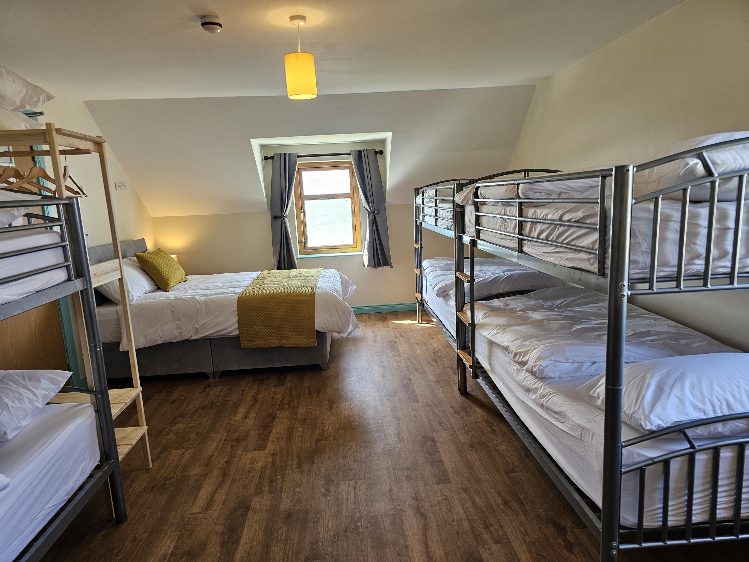 Room with four metal bunk beds, a window with curtains, a bed with a yellow blanket, and a small nightstand with a lamp.