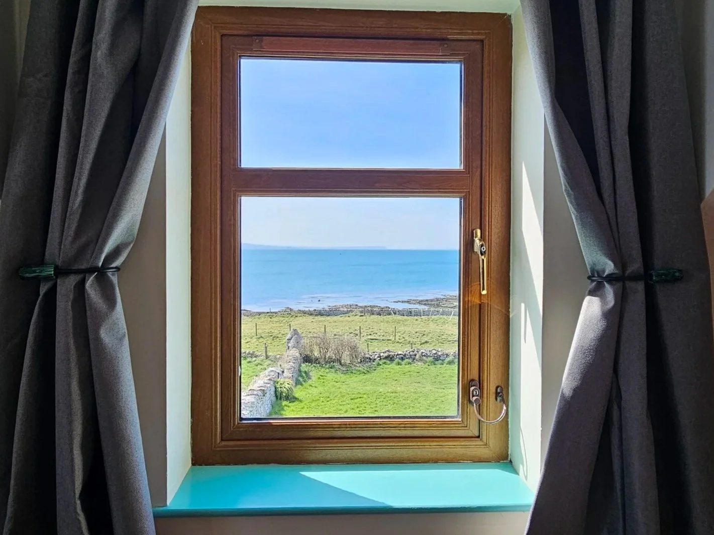 View through a window of the ocean, blue sky, grassy landscape, and rocks, with dark gray curtains on each side.