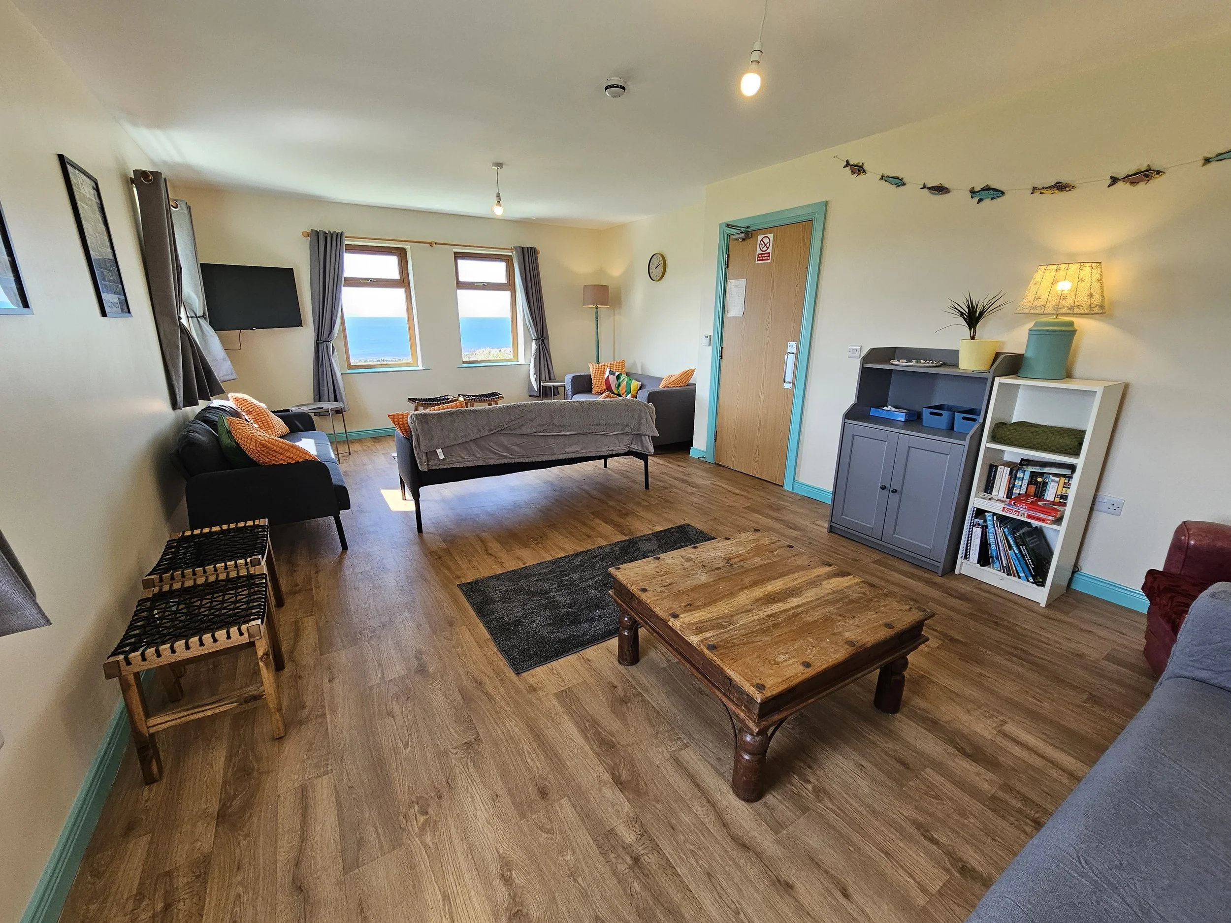 Living room with light-colored walls, wooden flooring, multiple windows with gray curtains, and furnishings including black and gray sofas with colorful pillows. There is a small black table, a wooden coffee table, a bookshelf, a gray cabinet with bl