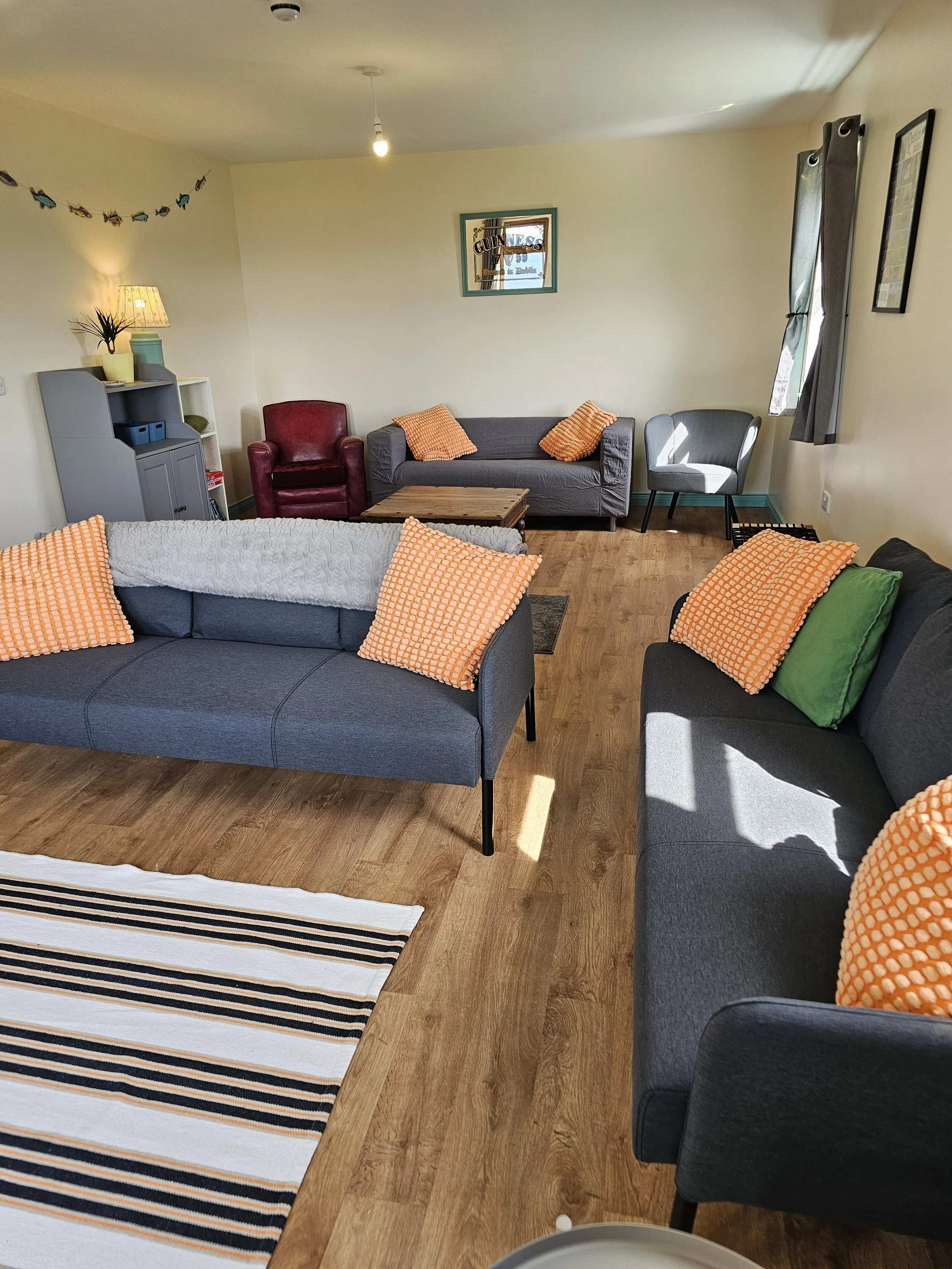 Living room with gray sofa, black and green pillows, orange pillows, a red armchair, a small wooden coffee table, a gray shelving unit with lamp, potted plant, and decorative items, a window with closed curtains on the right, framed pictures on the w
