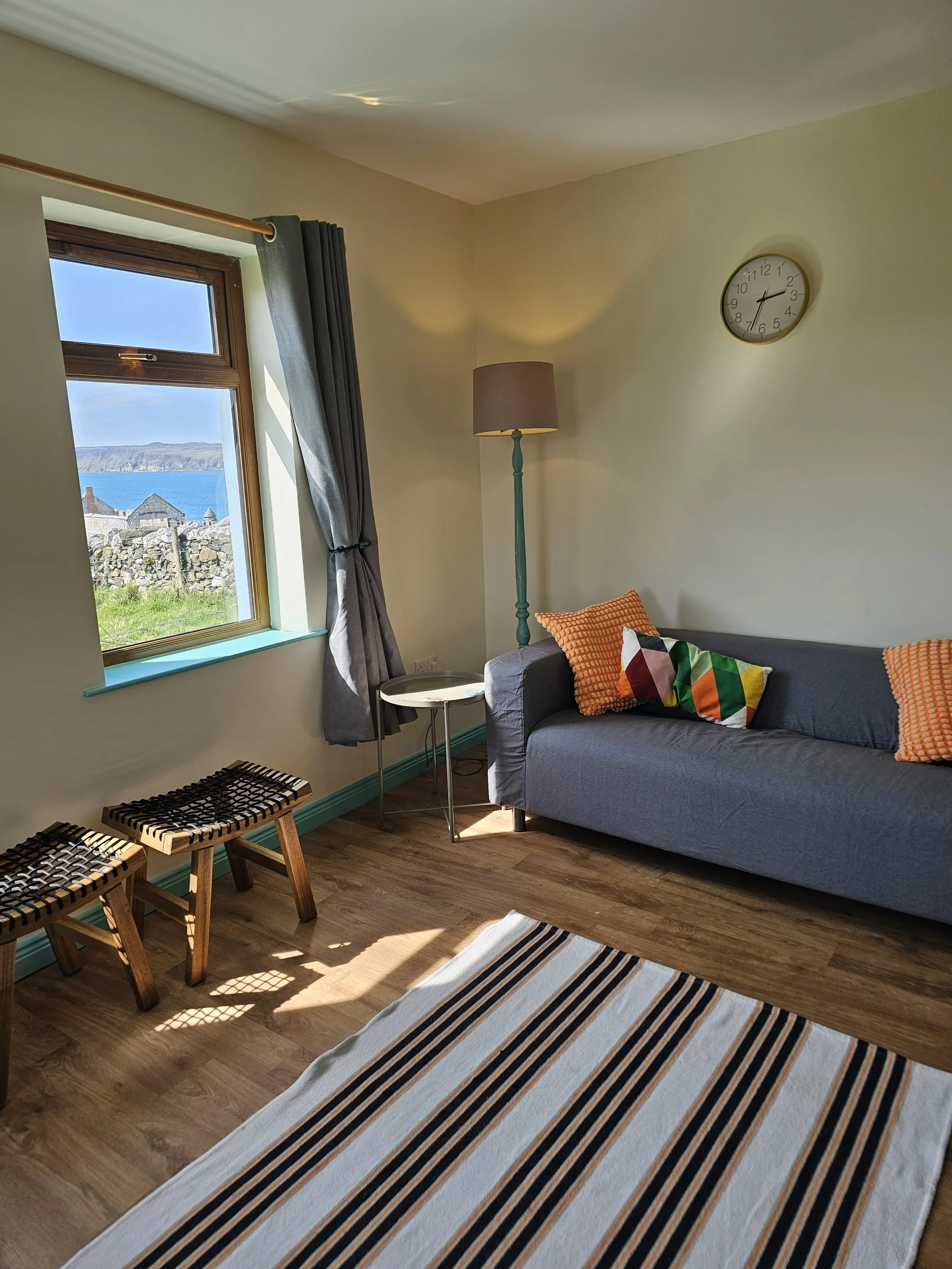 Living room with a window showing a view of the ocean and cliffs, a gray sofa with colorful pillows, a striped rug, a tall lamp, and two wooden stools.