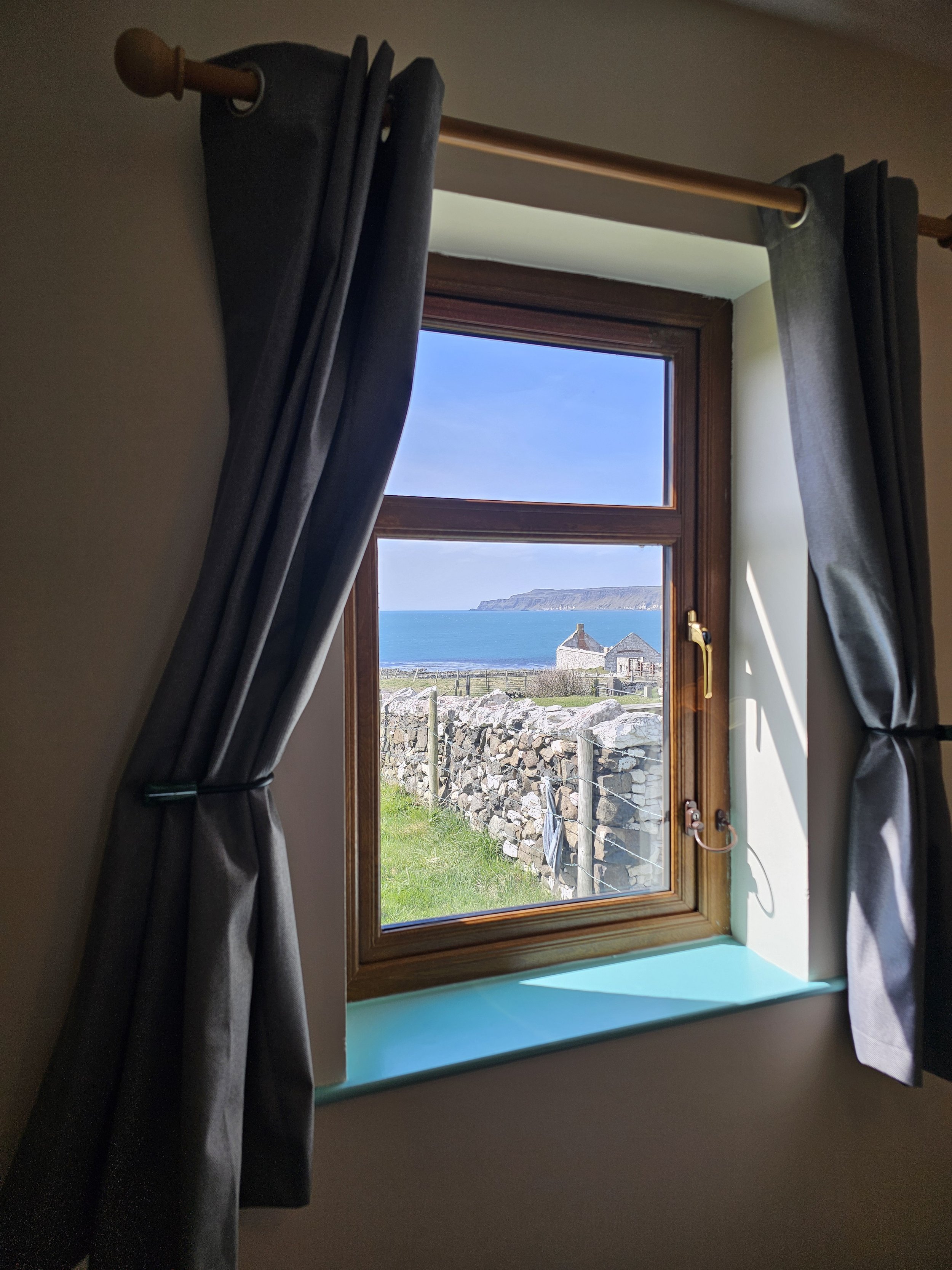 A view through a window showing a coastline with a hill in the distance, a stone wall outside, and a white building.