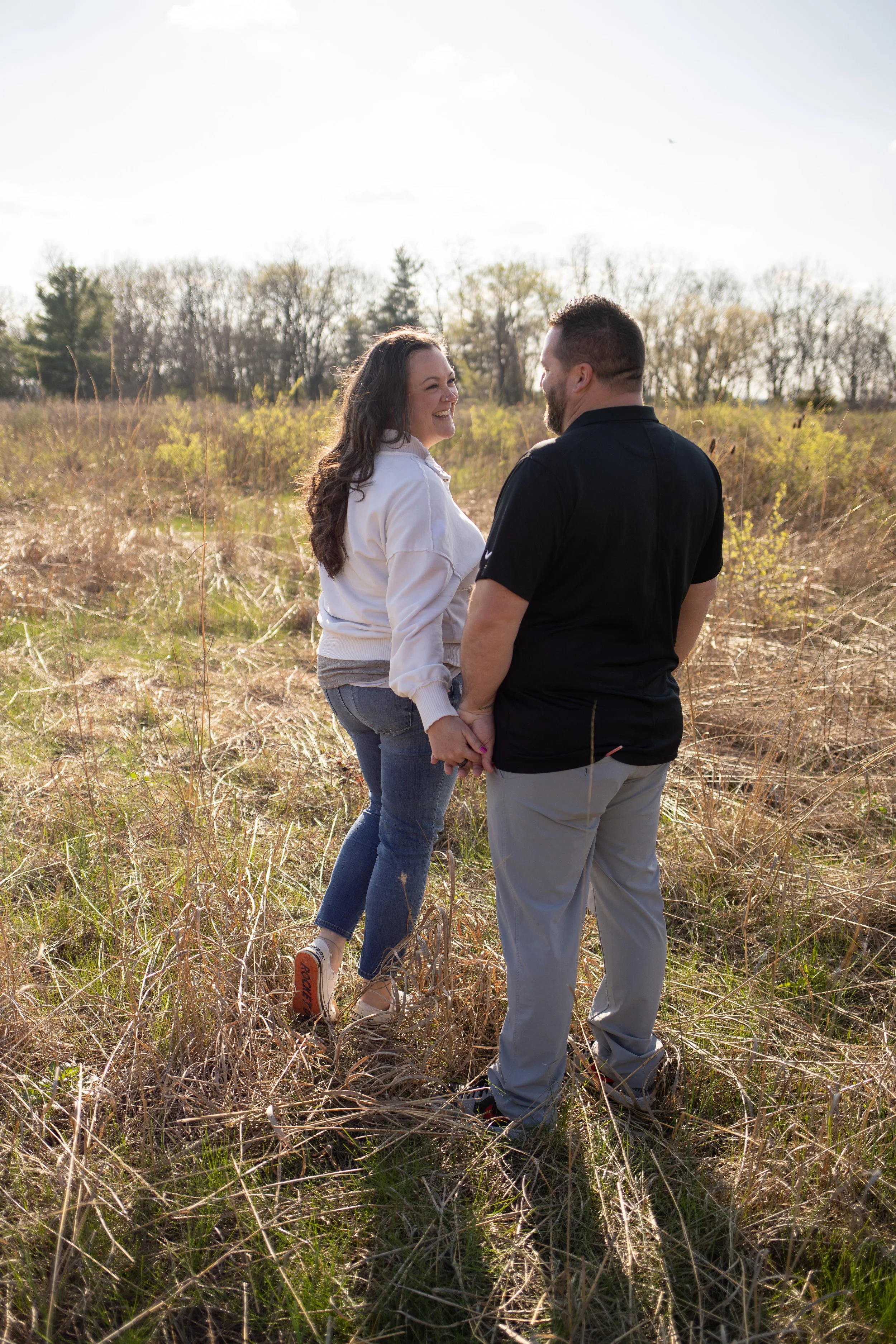 A couple holding hands and smiling at each other outdoors in a field with tall grass and trees in the background, during daylight.