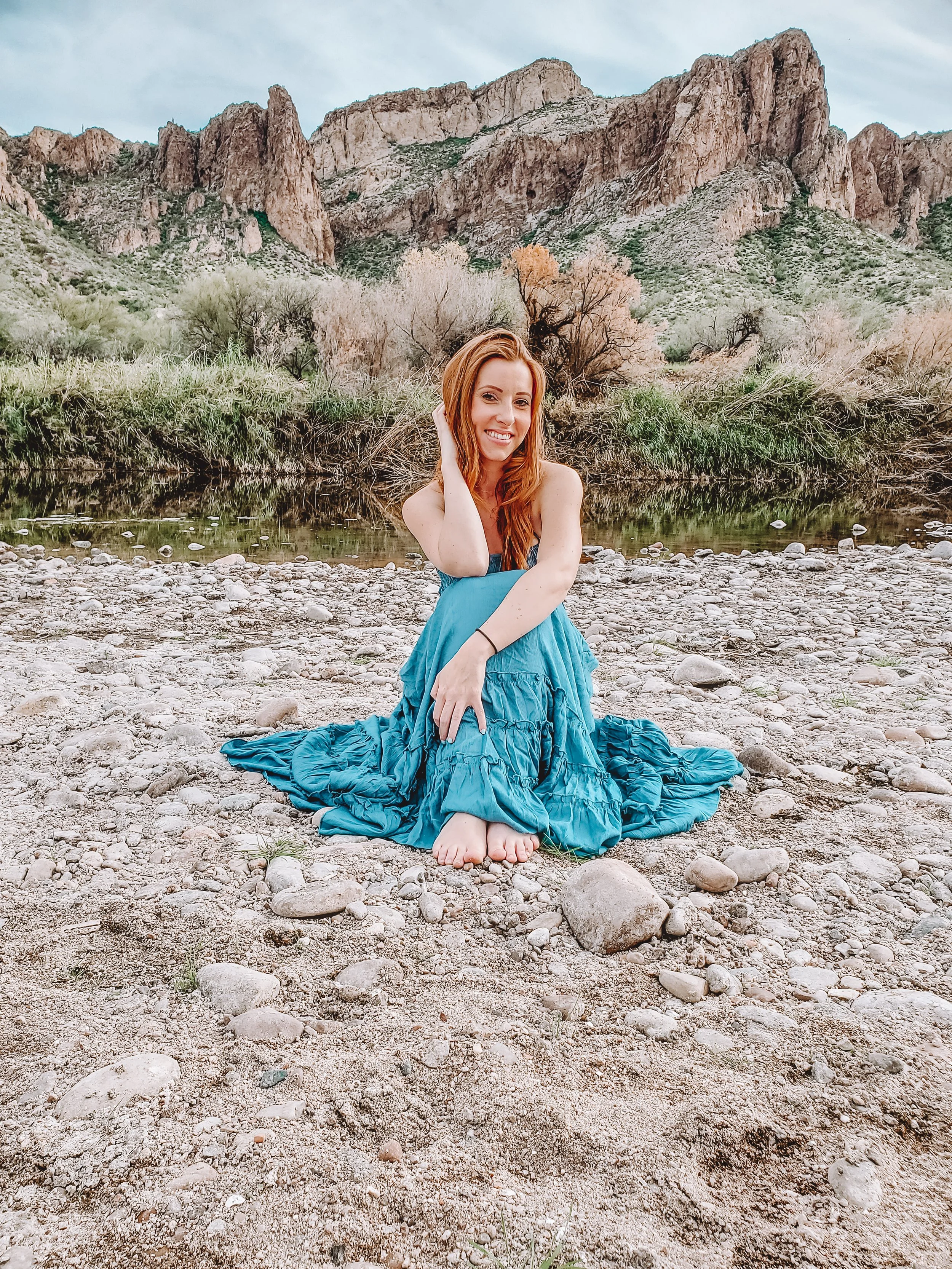 A smiling woman with red hair in a blue dress sitting on a rocky riverbank with mountains and trees in the background.