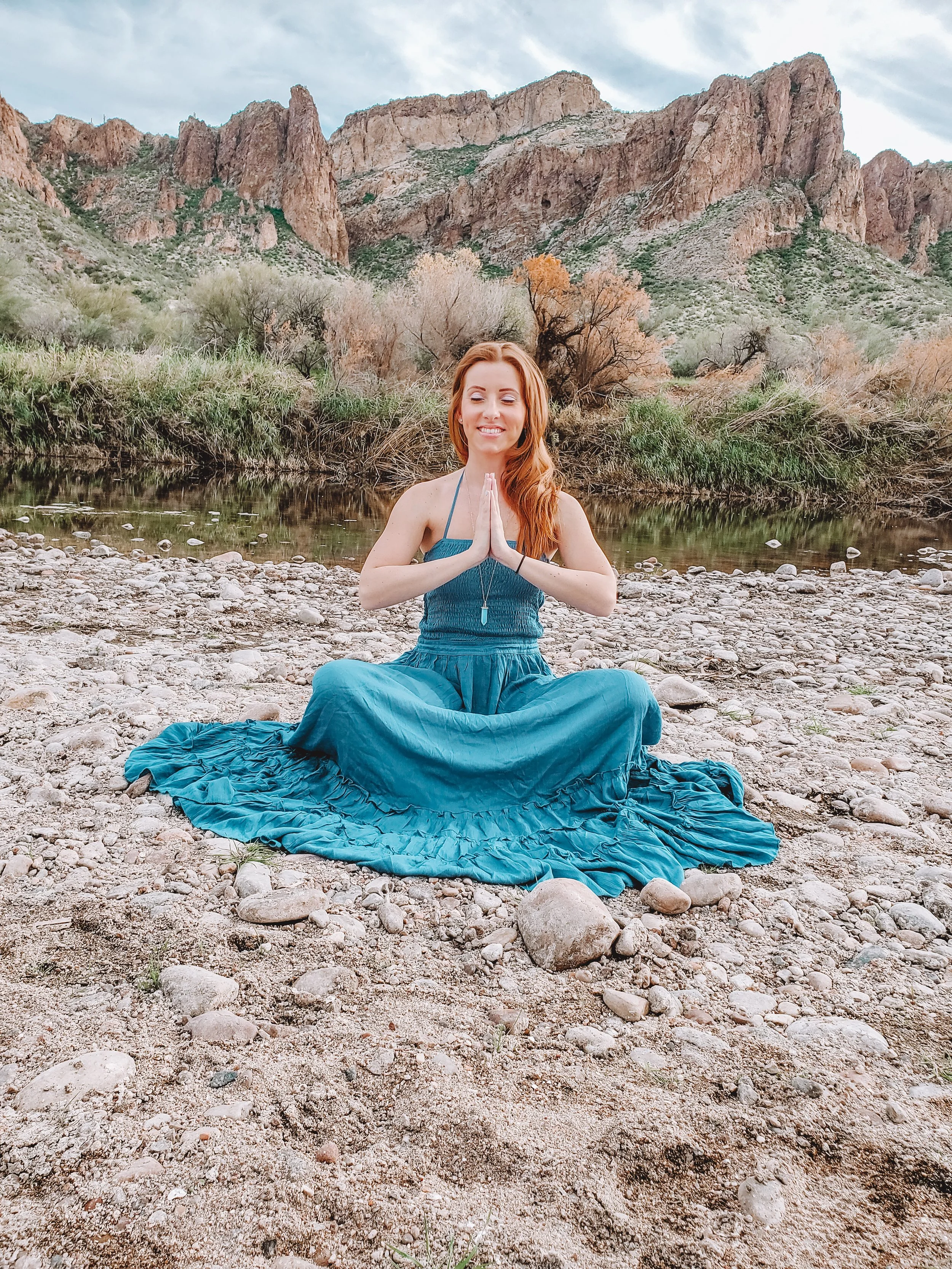A woman with long red hair is sitting cross-legged on a rocky riverbank. She has her hands in a prayer position in front of her chest and is smiling with her eyes closed. She is wearing a teal dress and a pendant necklace. In the background, there is a calm river, green bushes, and tall, rugged mountains under a partly cloudy sky.