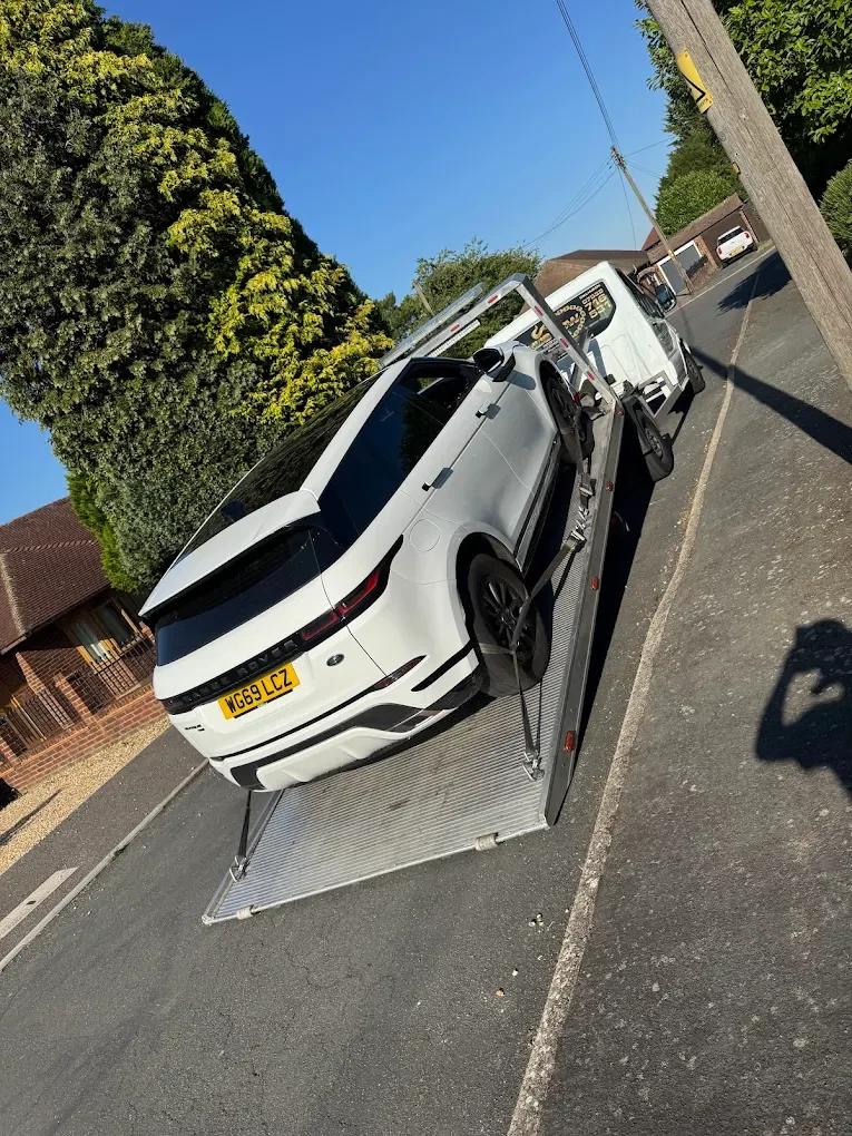 White Range Rover Evoque on a flatbed tow truck parked on side of street with houses, trees, utility poles, and a clear blue sky in the background.