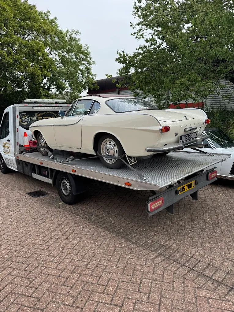 A classic white vintage car is on a flatbed tow truck, parked on a brick driveway with trees and a building in the background.
