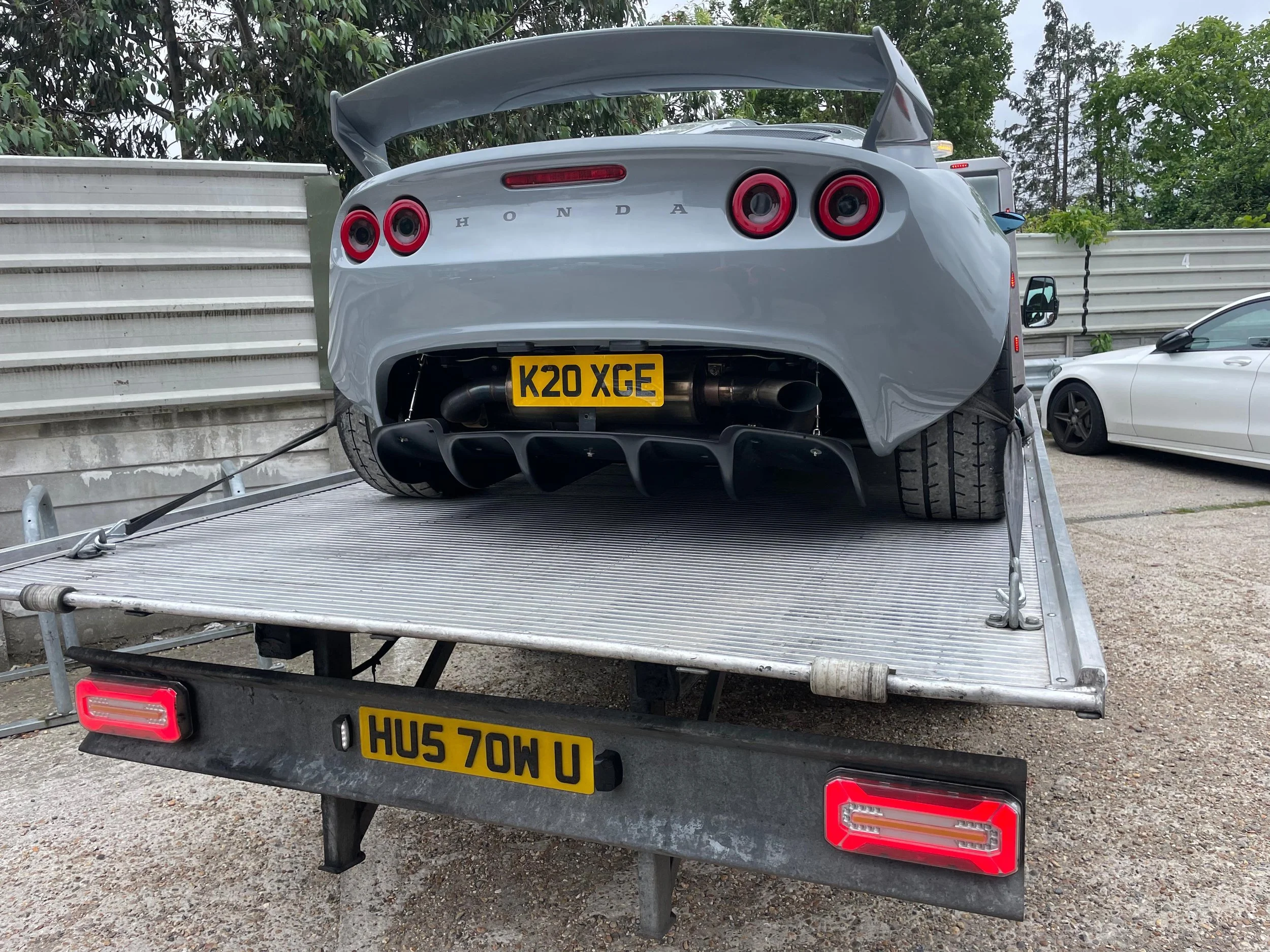 A silver sports car with a large rear wing, distinctive round taillights, and a yellow UK license plate, secured on a flatbed trailer with a metal ramp, parked outdoors against a backdrop of trees and a gray sky.