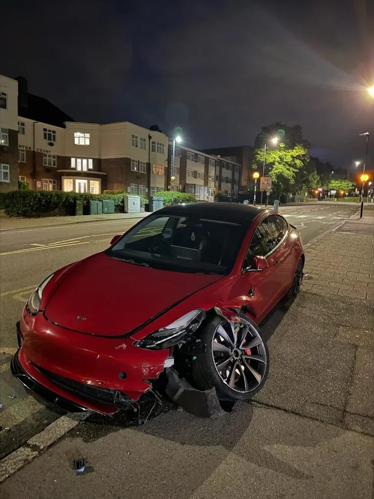 Red car with front end damage and flat tire parked on the side of a street at night.
