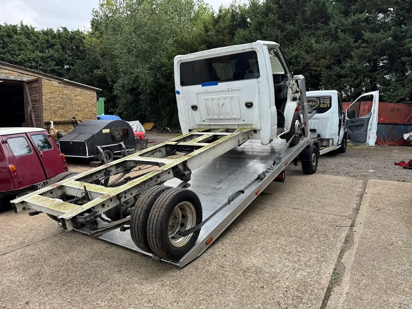 A flatbed tow truck carrying a disassembled white truck cab with no wheels, parked on a gravel lot with trees and various vehicles in the background.