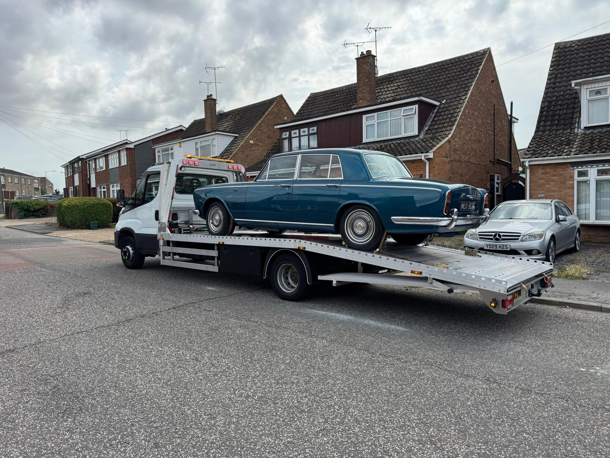 A classic blue 1960s car is loaded onto a flatbed tow truck on a residential street. The street is lined with brick and siding houses with pitched roofs and chimneys, under a cloudy sky.