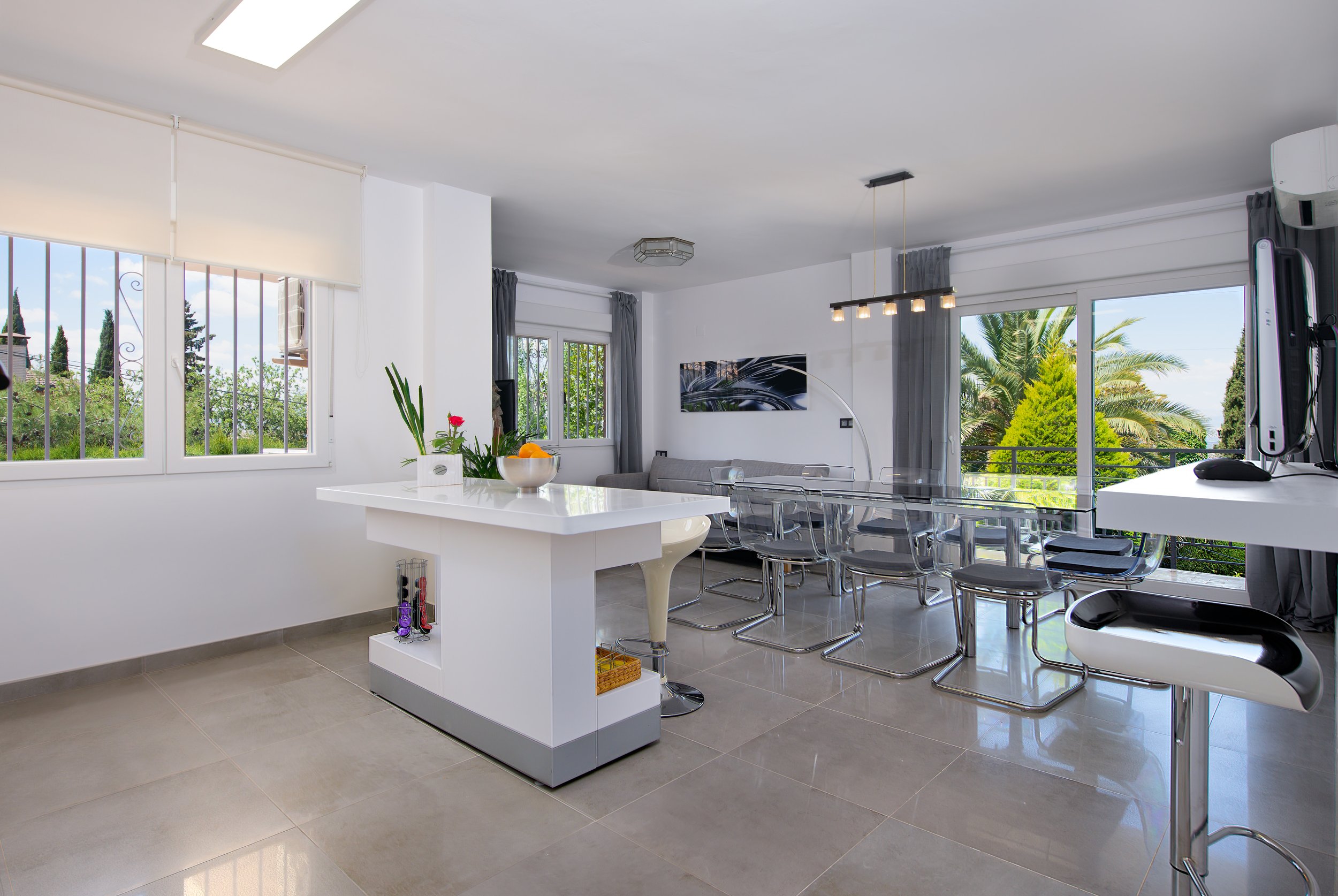 Sala de estar moderna con ventanas grandes, vista a plantas y árboles, mesa blanca con flores y fruta, comedor con sillas transparentes, televisor, cortinas grises y luces colgantes.