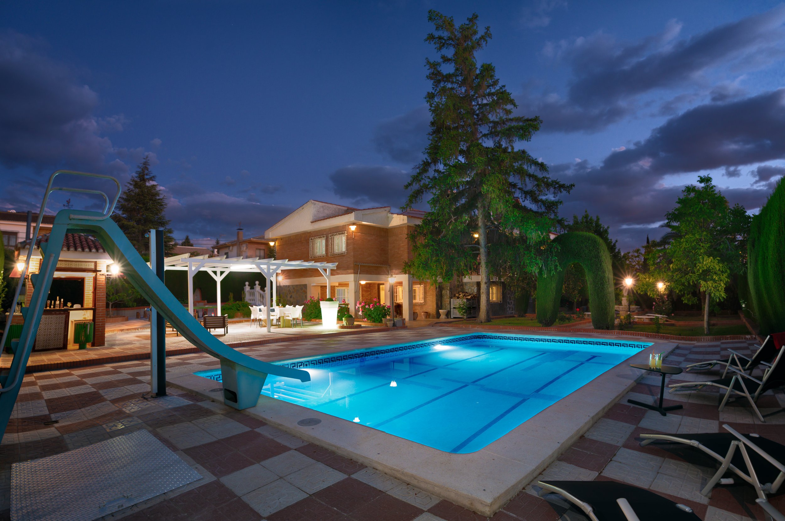 Piscina iluminada por la noche en un jardín con palmeras, casas y cielo nublado