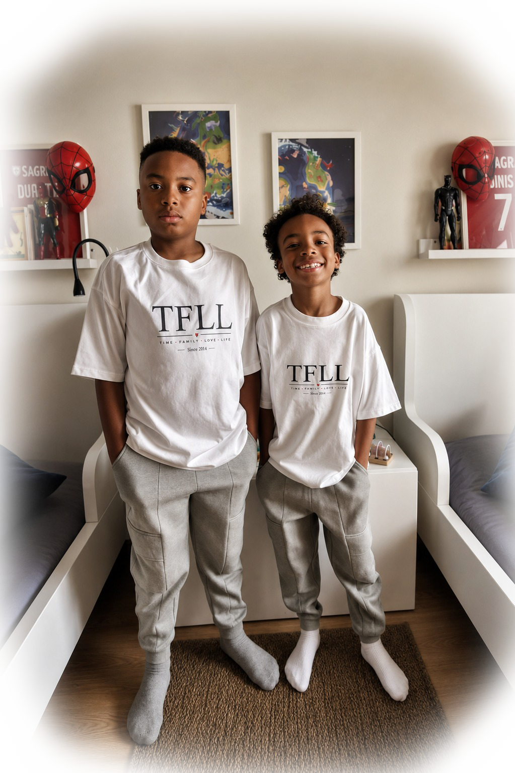 Dois meninos jovens estão de pé no quarto, vestindo camisetas brancas e calças cinza, com meias cinza e brancas, sorrindo e olhando para a câmera.