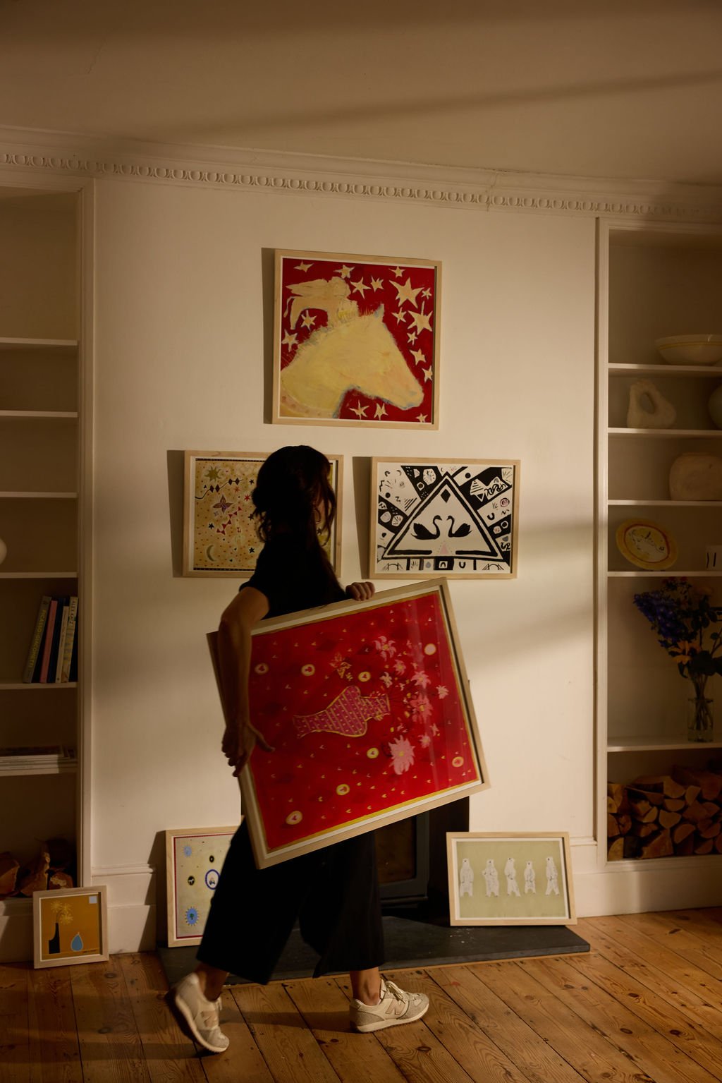 A woman in black pants and sneakers standing in a living room holding a framed artwork with a red background and a fish design. The room has wooden floors, white walls, decorative crown molding, and built-in bookshelves with various items and artwork displayed on the wall and leaning against the fireplace.