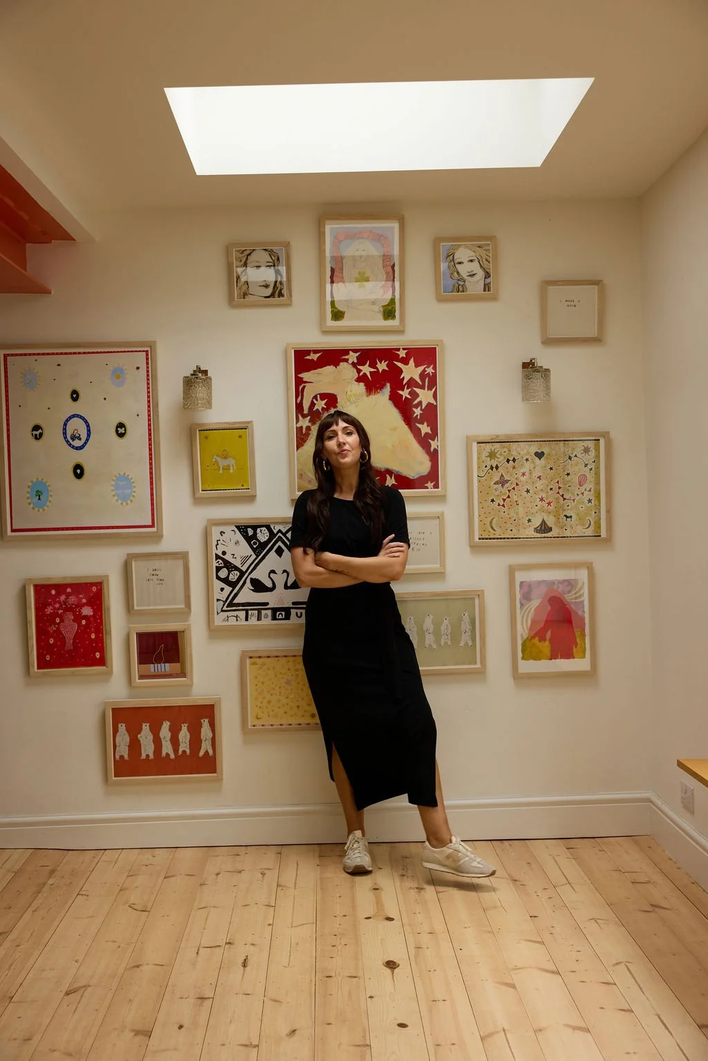 A woman standing with crossed arms in an art gallery, surrounded by various framed artworks on a white wall, with a wooden floor and a skylight above.