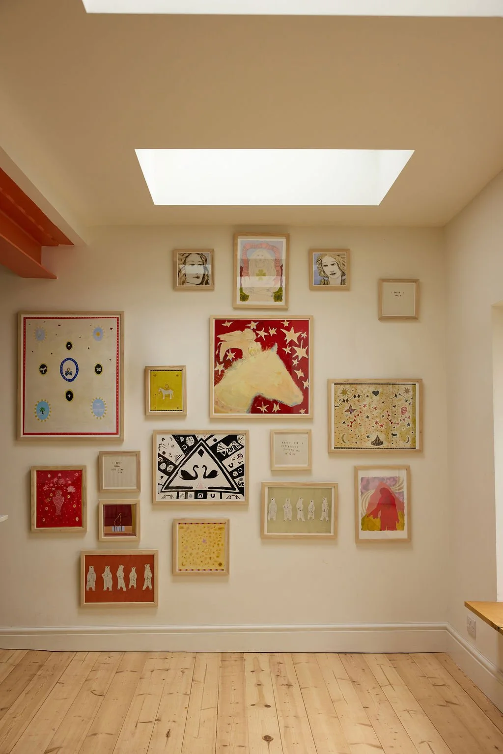 A gallery wall with framed colorful artworks and illustrations, featuring a prominent central piece of a horse's head with a red background, above a light wooden floor and under a ceiling with a skylight.