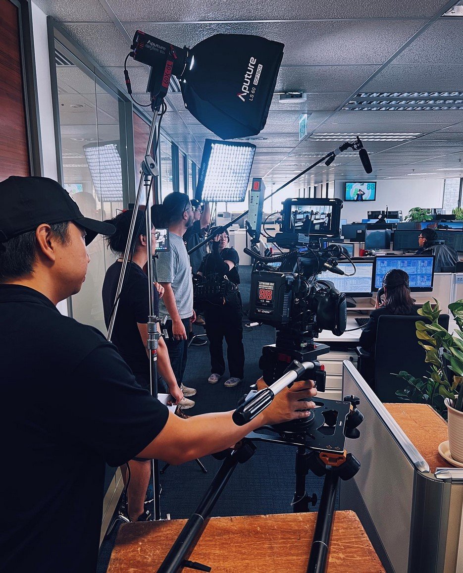 Film crew filming inside an office, with a camera on a tripod, lighting equipment, and people working at desks in the background.