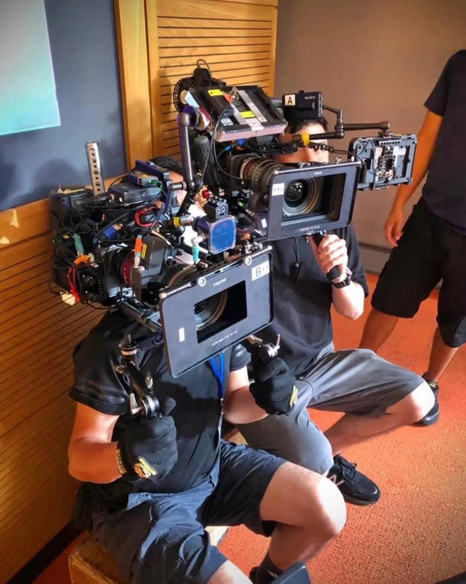 Two people are holding large camera rigs with attached monitors and equipment in a room with wooden paneling and orange carpet. One person is kneeling while holding the camera rig, wearing gloves, a black t-shirt, and shorts. The other person is standing, wearing black clothing.