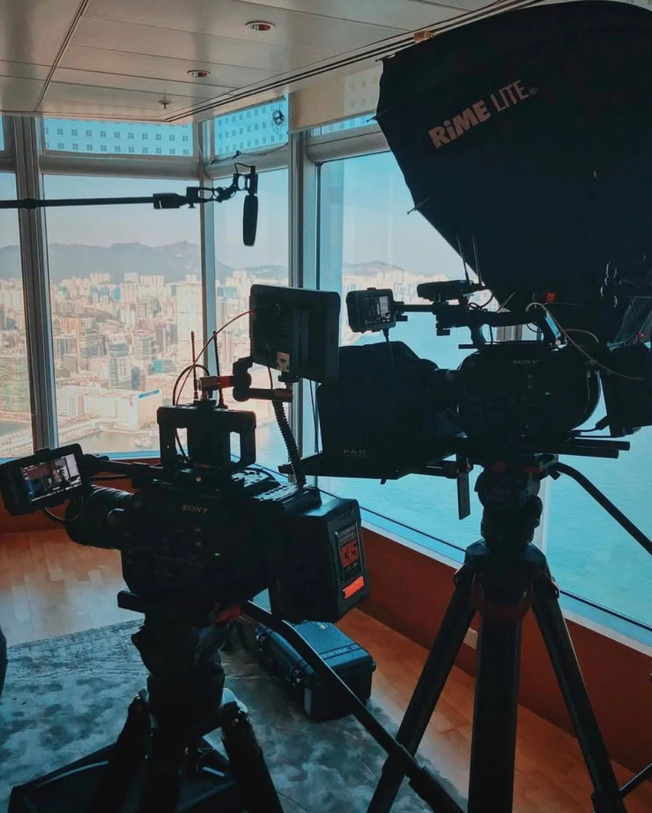 Inside a high-rise building with a large window showing a cityscape, camera equipment and lighting gear set up for a video or photo shoot. The scene includes professional cameras, tripods, a large umbrella light, and a boom microphone.