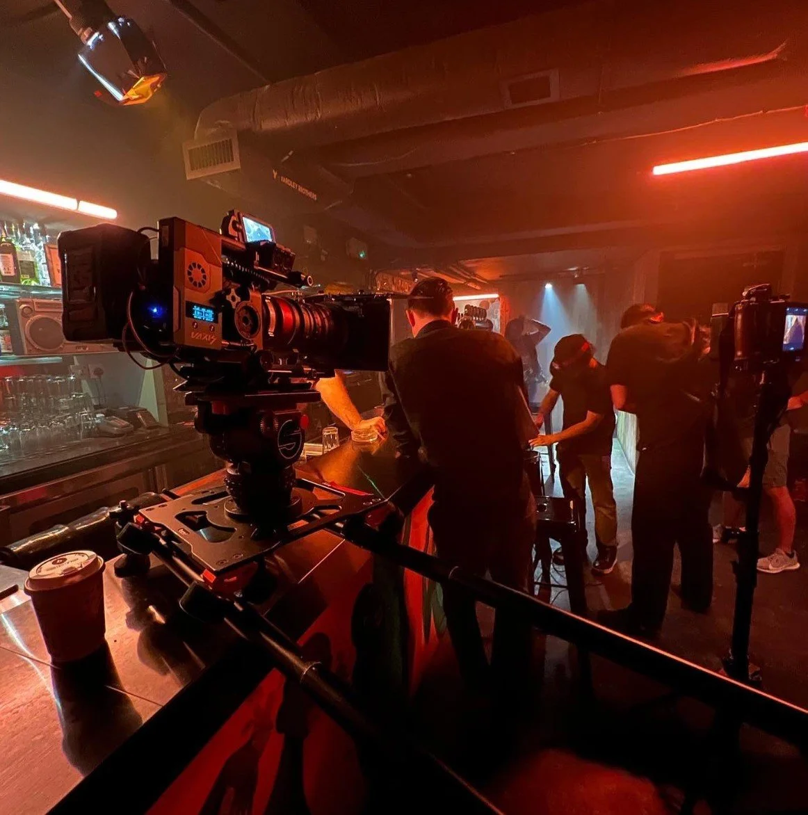 A film crew filming inside a nightclub with red and blue lighting, a camera, and crew members working behind the scenes.