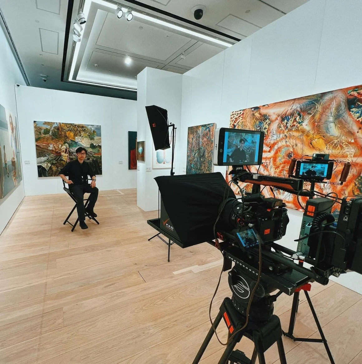 A person being filmed in an art gallery with large colorful paintings, using professional camera equipment and lighting.