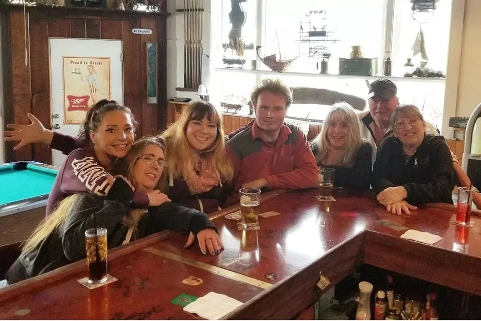 A group of six people sitting around a bar, smiling and enjoying drinks in a cozy, dimly-lit pub with a pool table nearby.