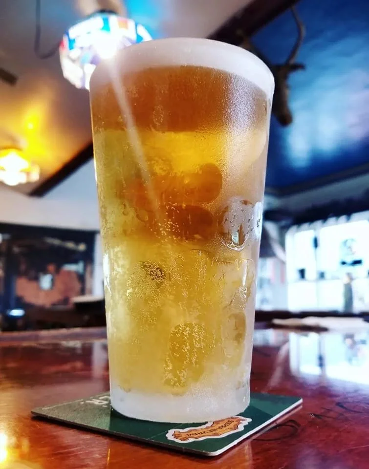 A tall glass of a chilled, clear beverage with ice cubes and a slice of lemon, placed on a coaster on a wooden table in a bar or restaurant setting with colorful lights in the background.