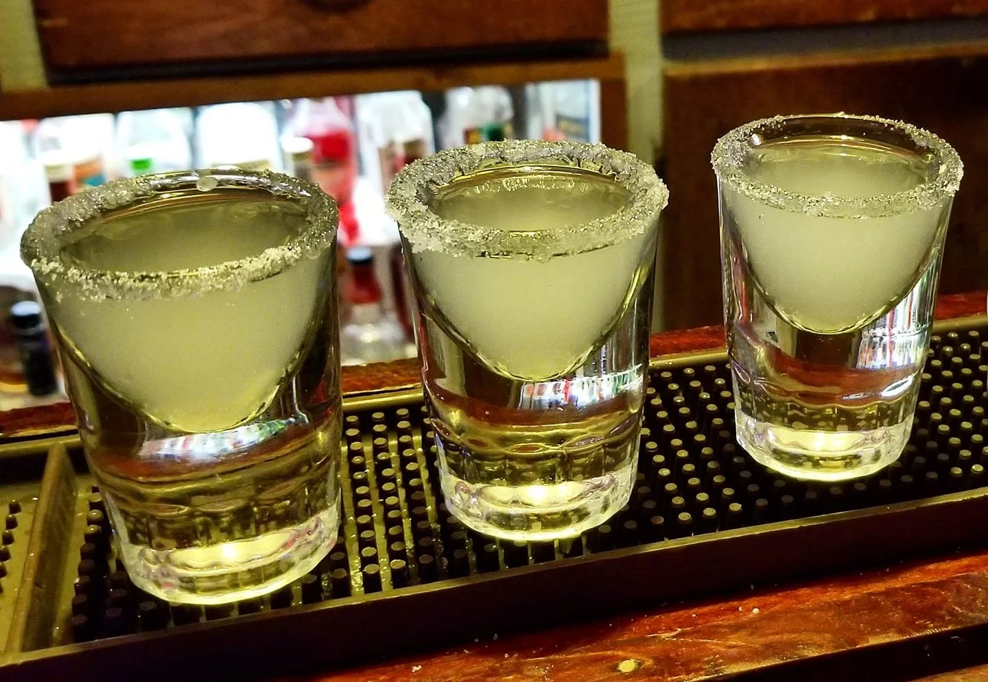 Three shot glasses filled with a clear yellow liquid, garnished with salted rims, on a bar mat surface.