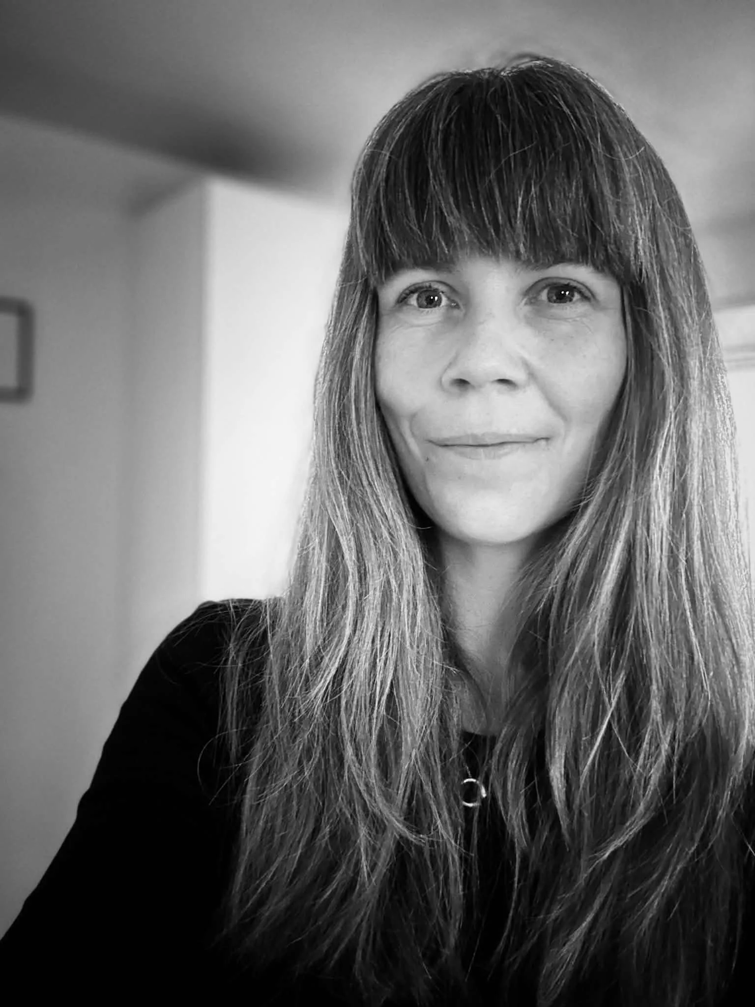 Sort-hvid portrætfotografi af en ung kvinde med langt, lige hår og stort smil, iført sort tøj, i et indendørs miljø.