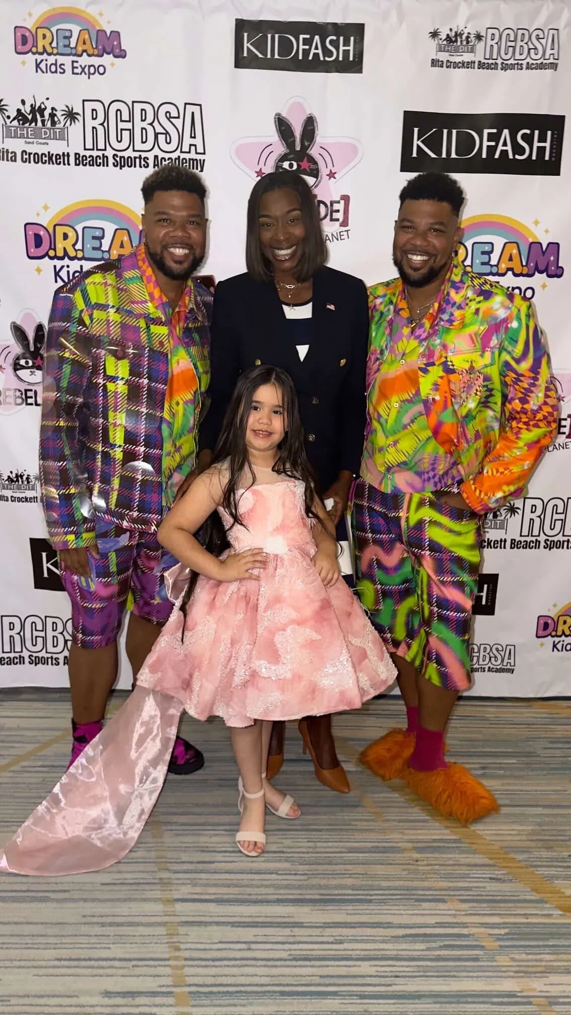 Three adults and one young girl posing at a kids expo with a step and repeat backdrop showing event and sponsor logos. The adults are wearing colorful, patterned outfits, and the girl is in a pink dress. One man is dressed as a furry orange creature.