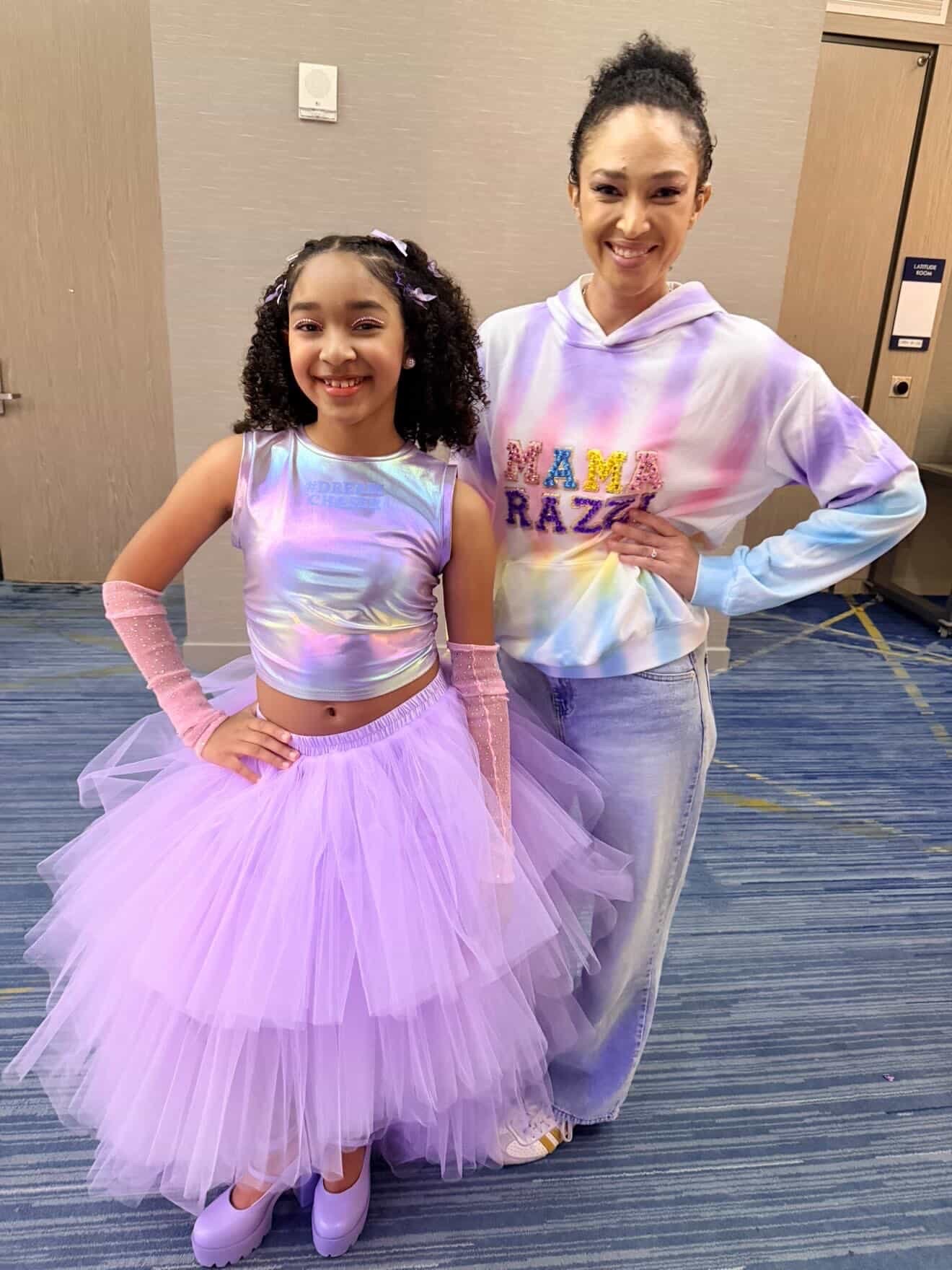 A young girl dressed as a ballerina in a lavender tutu and iridescent top, standing next to an adult woman wearing a pastel tie-dye hoodie with the words 'Mama Razz' embroidered on it. Both are smiling and posing indoors, in front of a beige wall and a door.