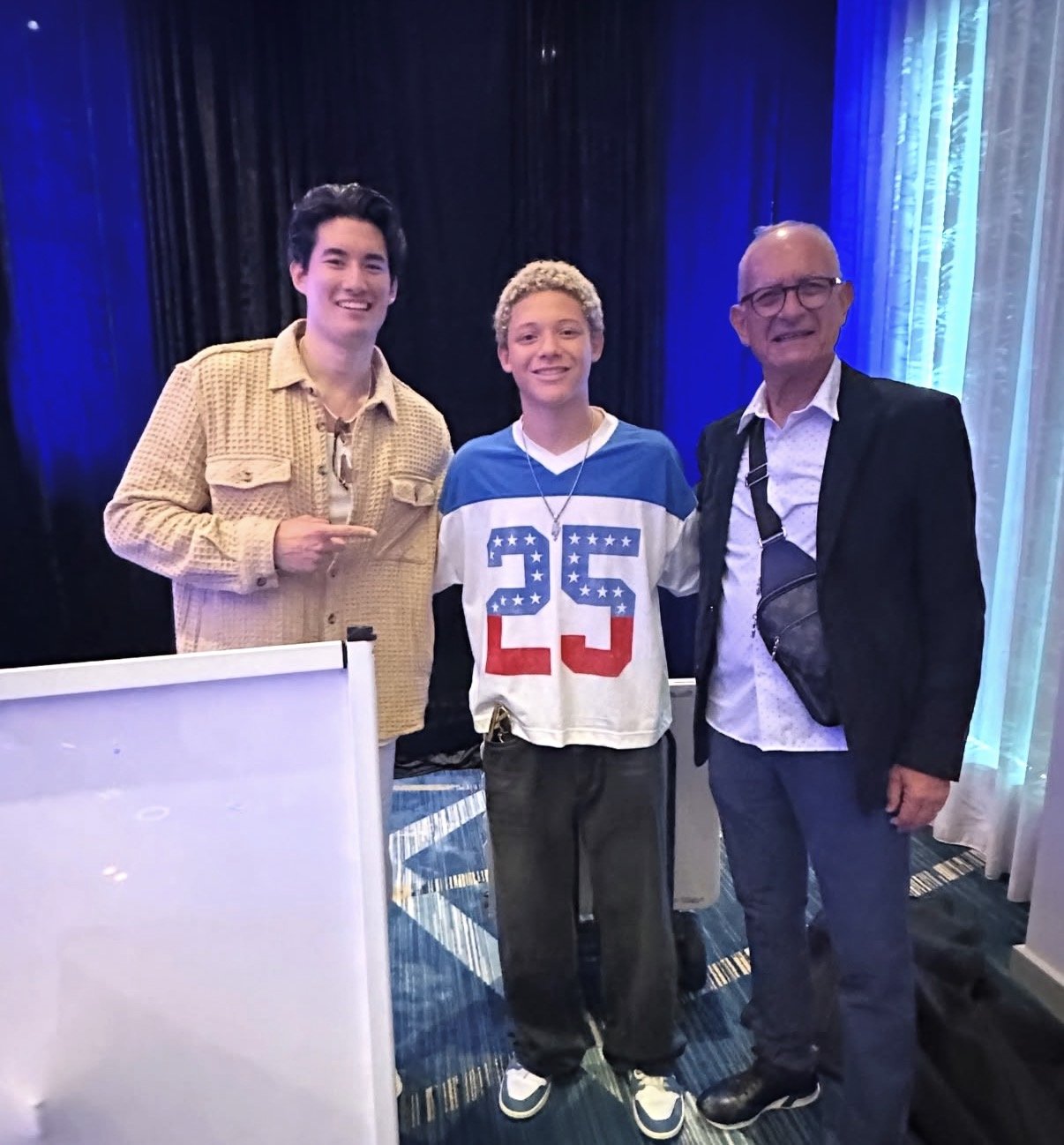 Three males standing together in a room with dark curtains and blue lighting. The person on the left is young, has dark hair, and is pointing with his right hand. The person in the middle is young, has curly blonde hair, and is wearing a jersey with the number 25. The person on the right is older, has glasses, and is dressed in a dark blazer over a white shirt, with a black strap across his chest.