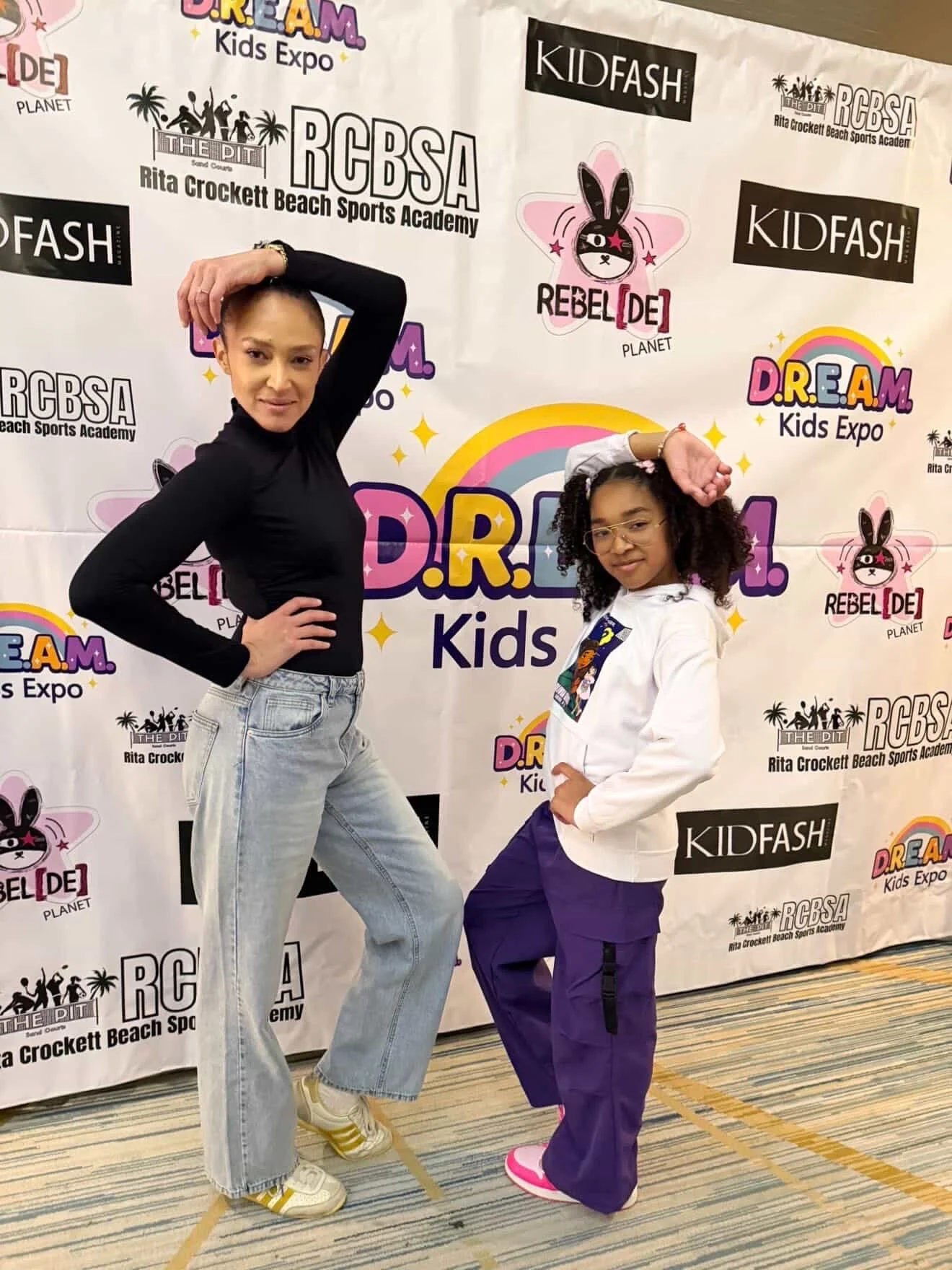 Two young women posing in front of a colorful event backdrop featuring logos for KidFash, D.R.E.A.M Kids Expo, and RCBS A at a kids expo event.
