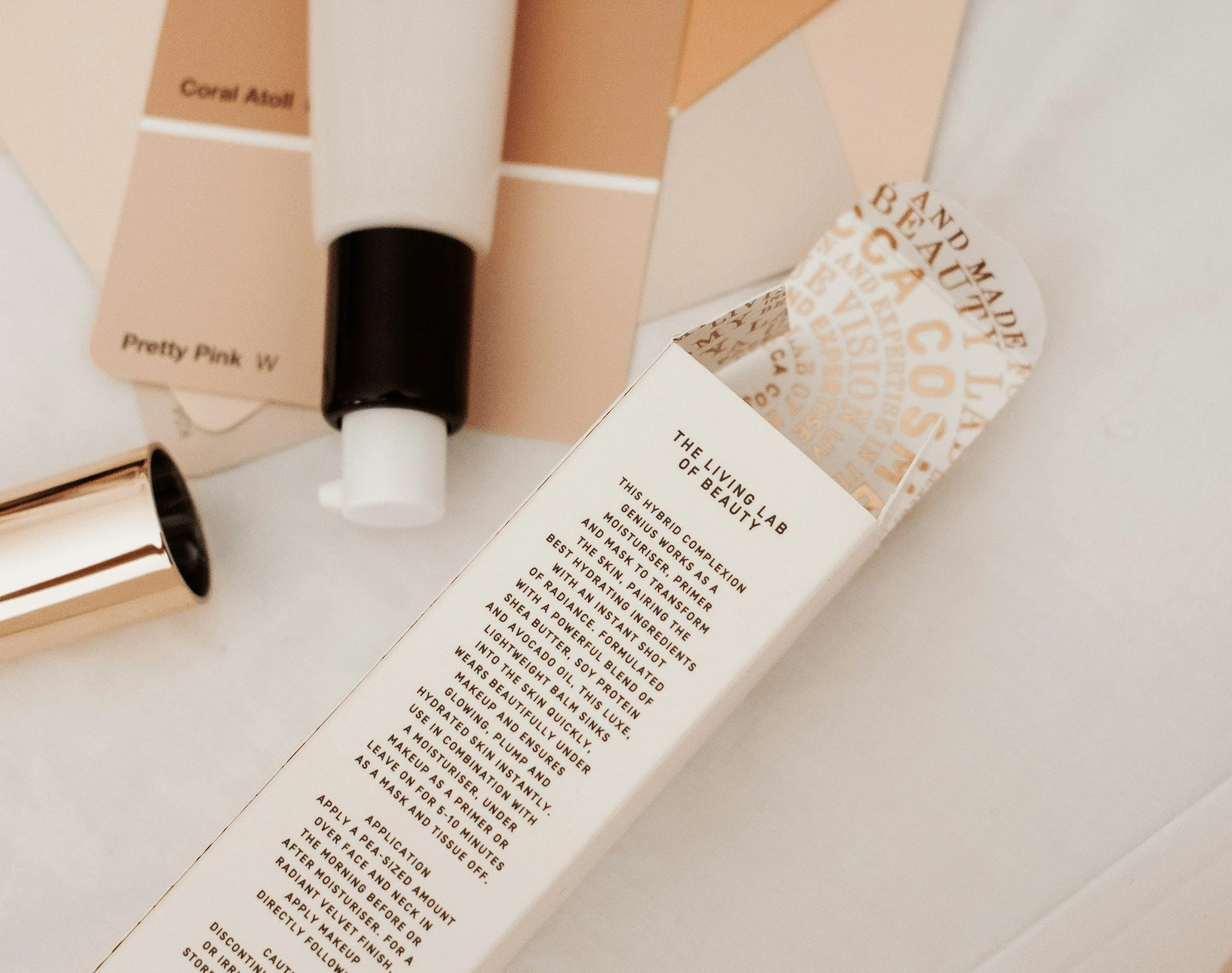 Close-up of skincare product packaging with text about a moisturizer, including shades labeled 'Coral Ash' and 'Pretty Pink W'.